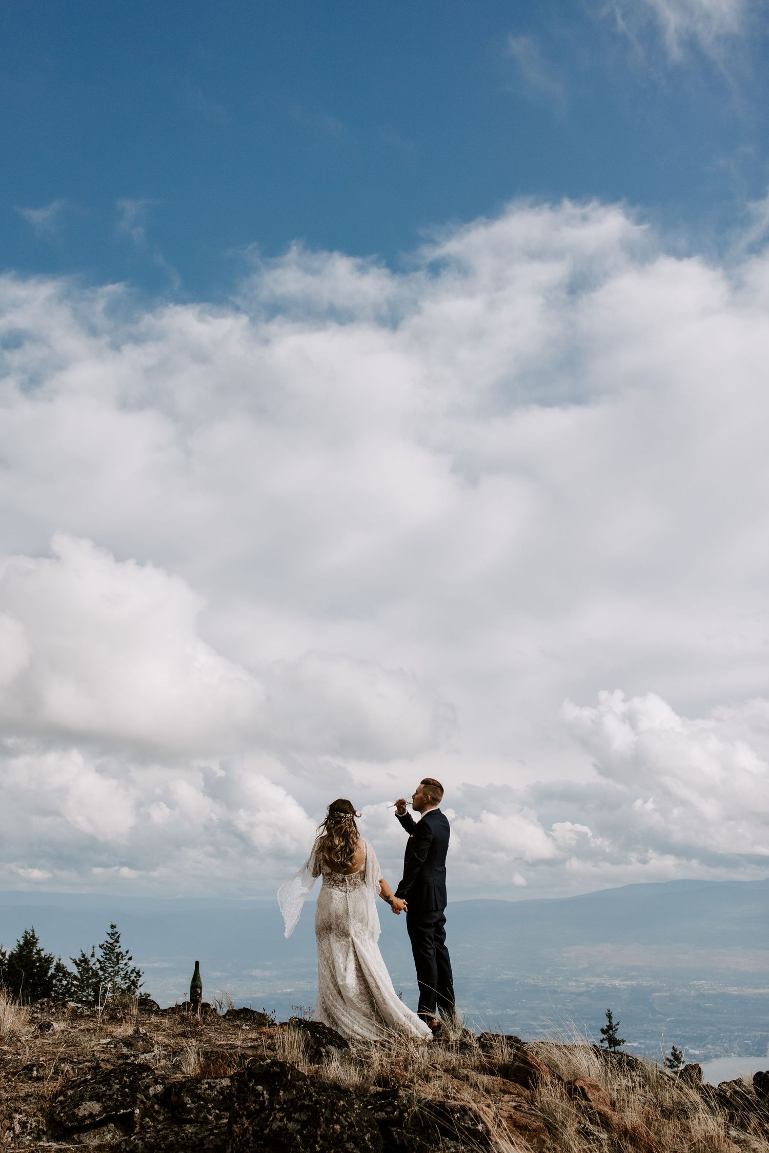 Silhouetted wedding couple stands on rocky mountain overlook against dramatic blue sky and clouds.