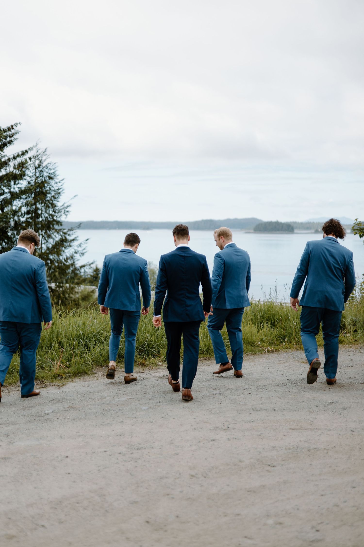 Five groomsmen in blue suits walk toward a scenic waterfront on a cloudy day, viewed from behind.