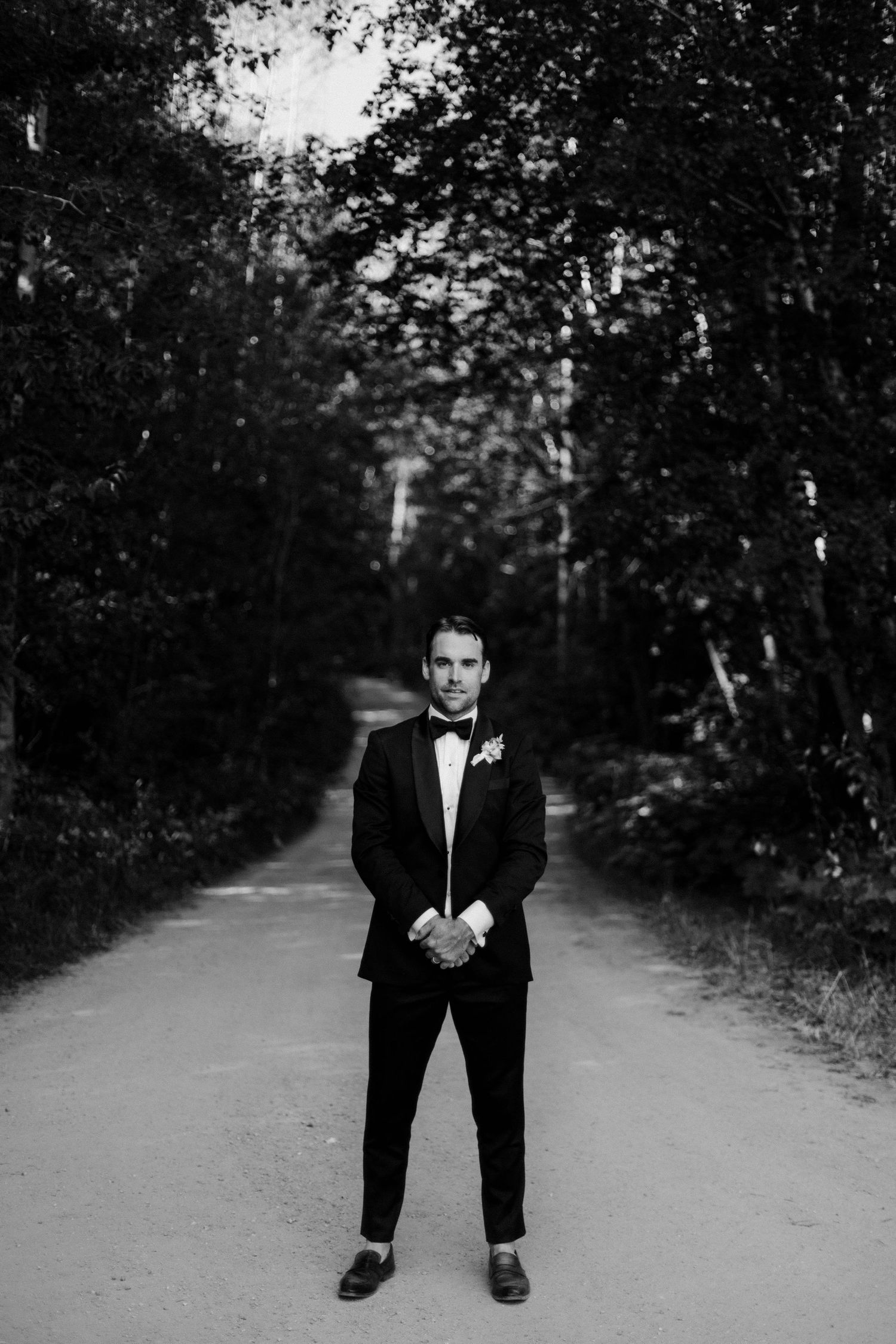 Groom in black tuxedo with boutonniere stands on a tree-lined path in a classic black and white wedding portrait.