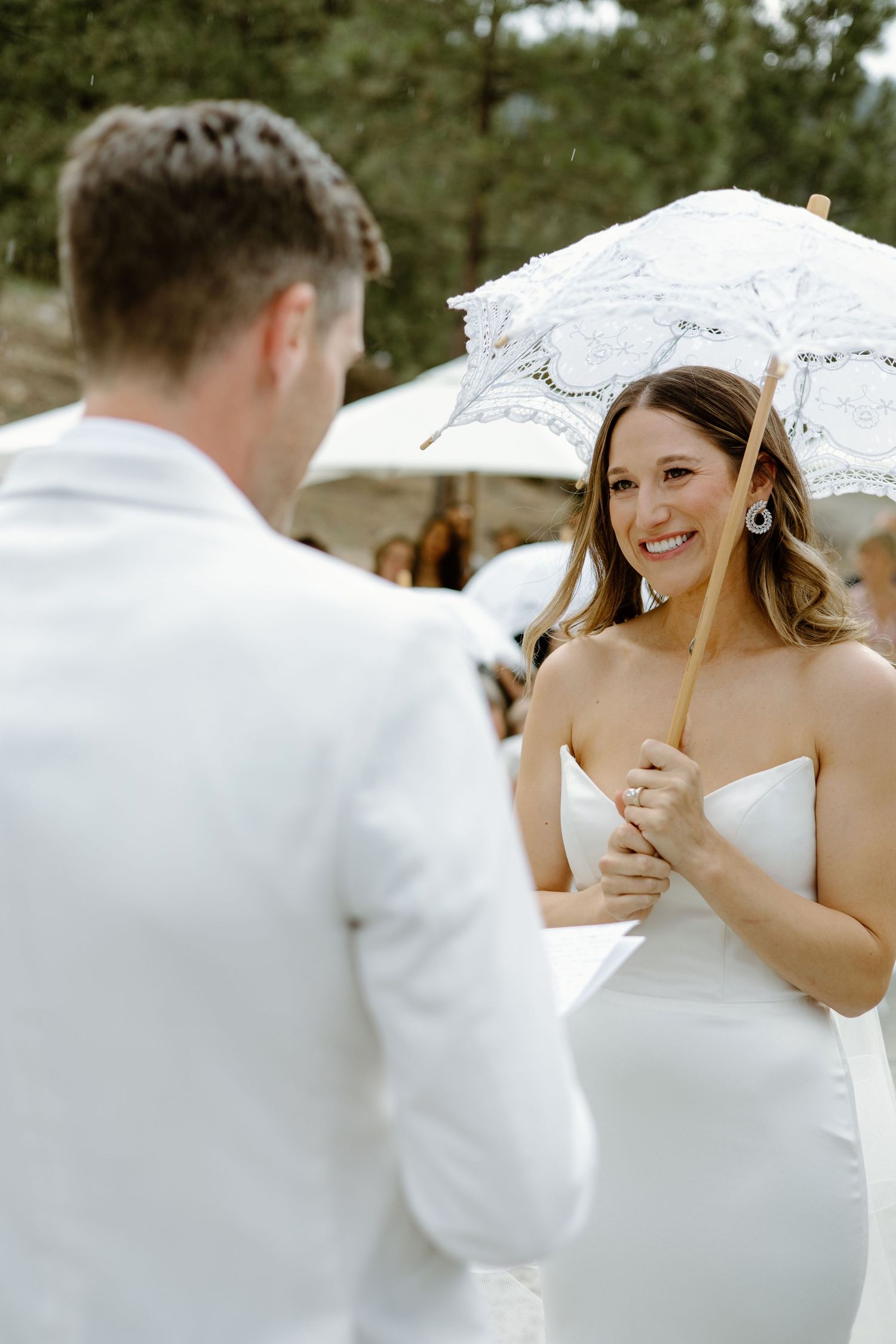Smiling bride in strapless gown holds white lace parasol facing groom in white suit at outdoor wedding ceremony.