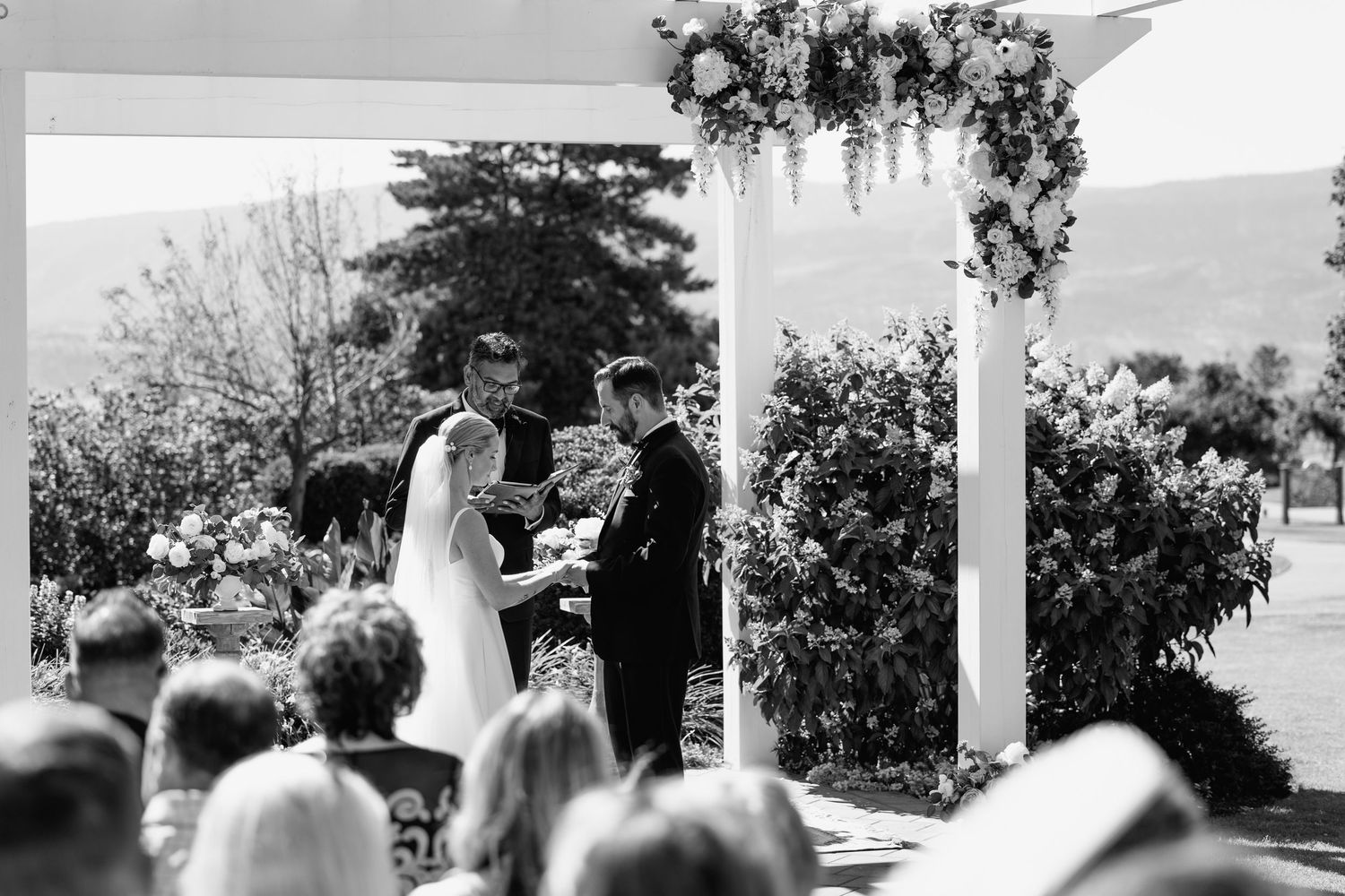 An outdoor wedding ceremony under a floral-adorned pergola with mountain views.