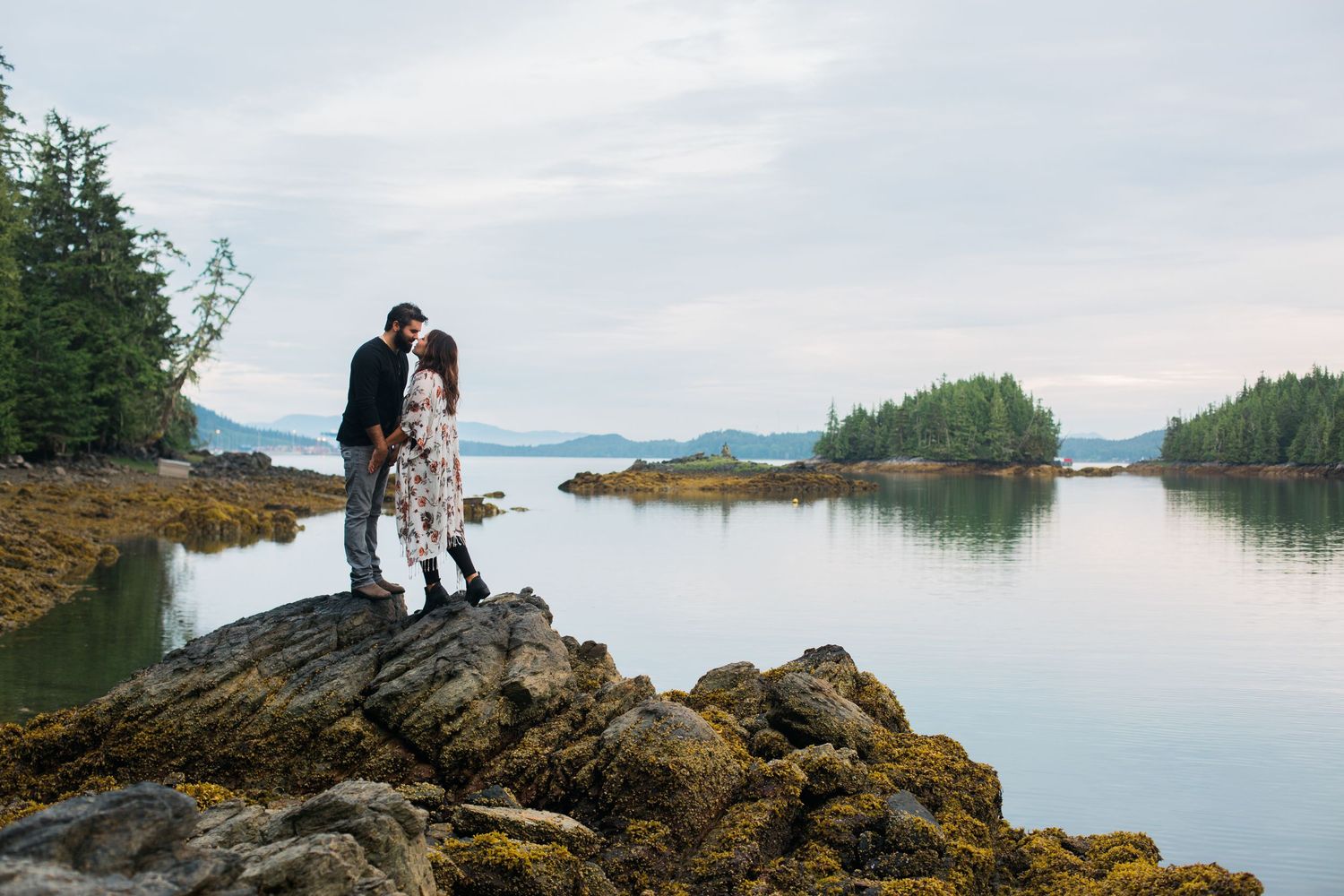 A couple poses romantically on coastal rocks with scenic water and islands in the background.