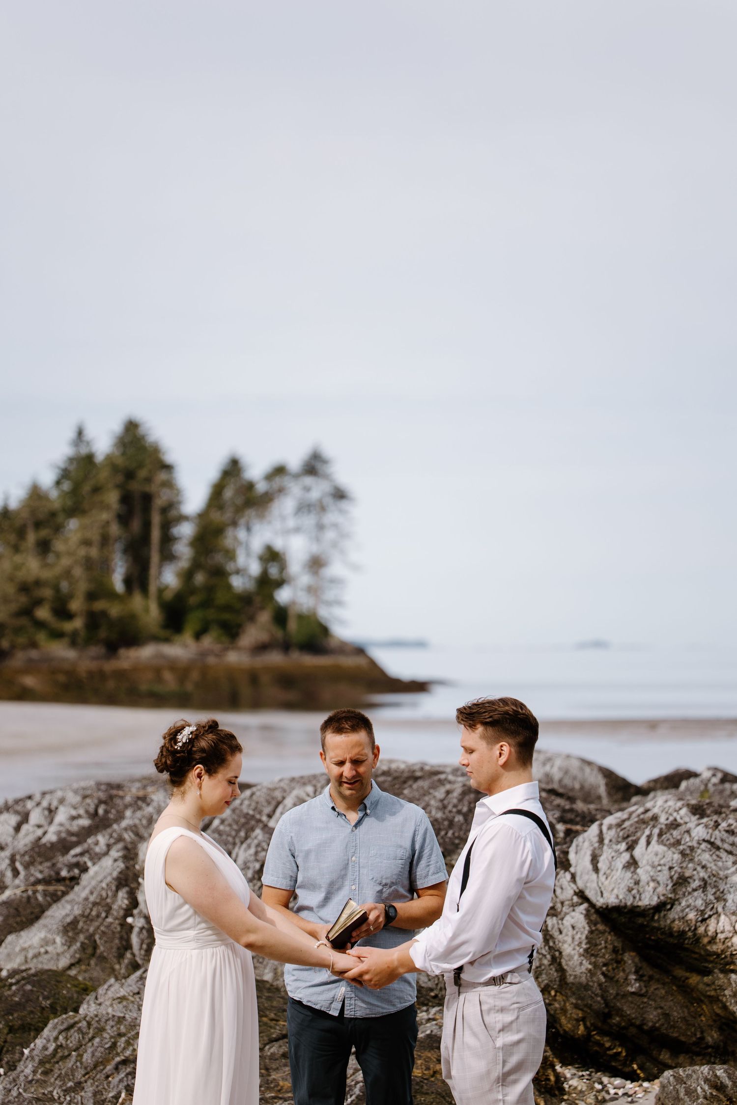 A couple shares an intimate wedding ceremony moment on a rocky Pacific Northwest beach with evergreen trees in background.