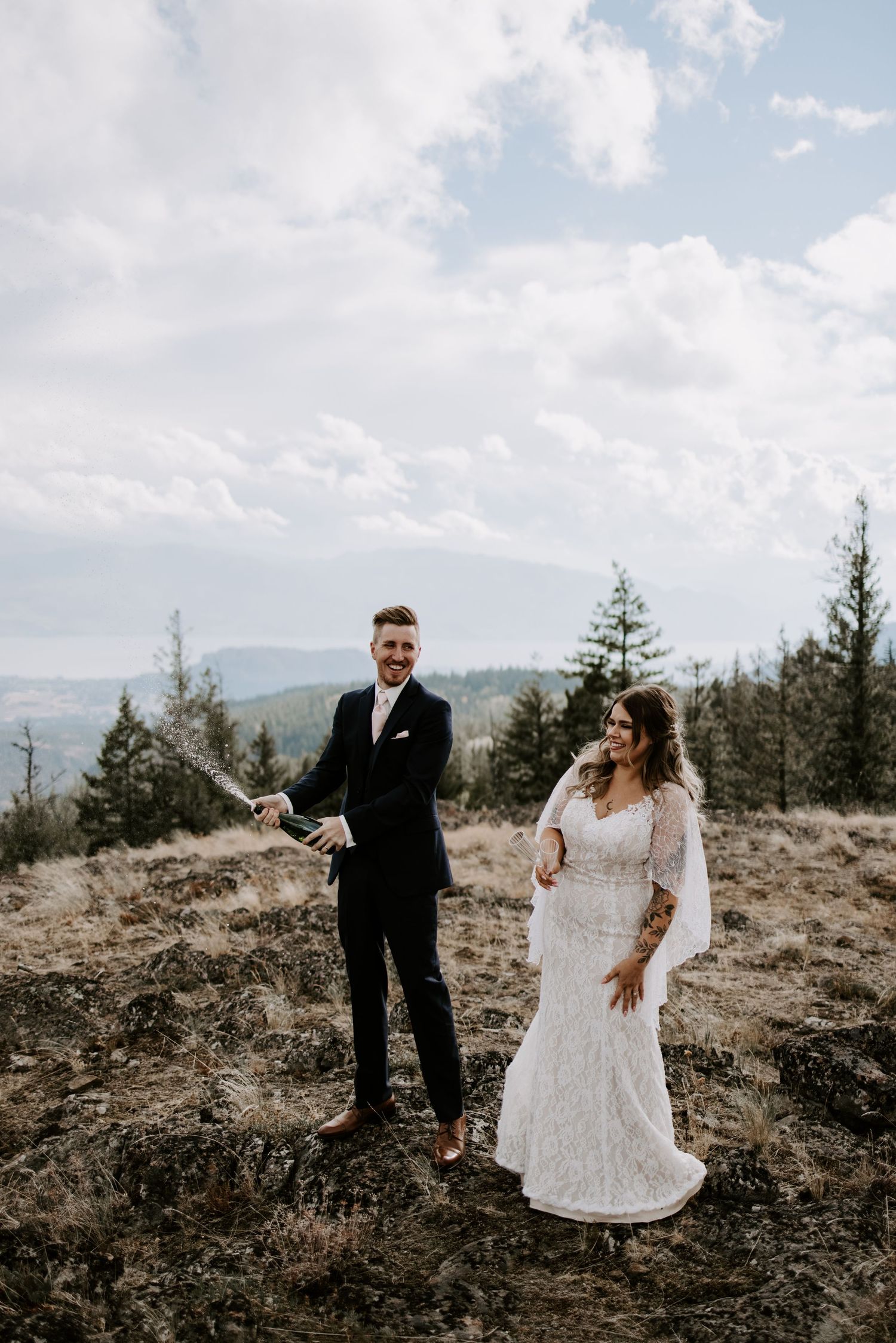 A couple in wedding attire shares joyful moments in a scenic mountain landscape with evergreen trees and cloudy skies.