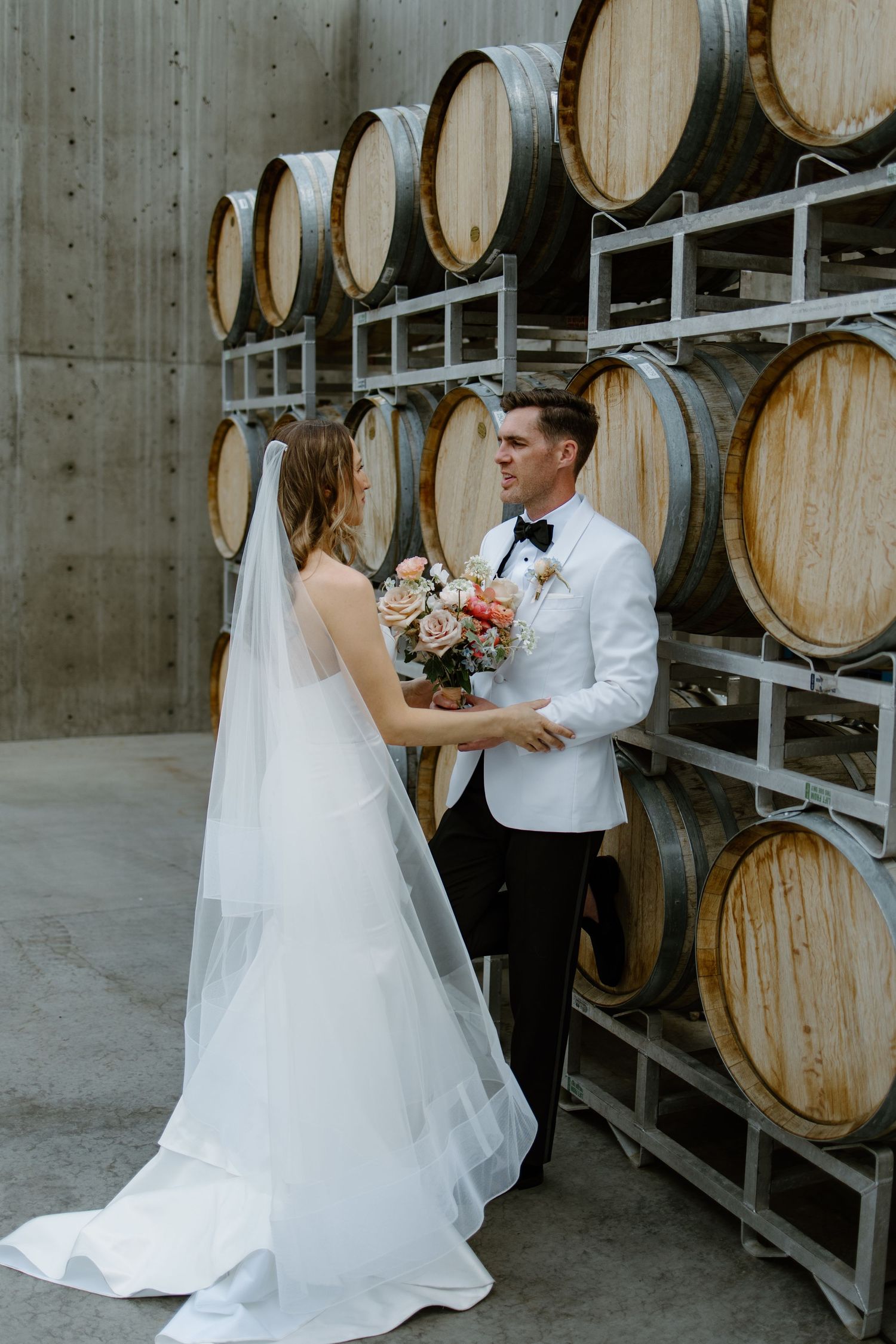 Bride and groom share a romantic moment holding a bouquet in front of oak wine barrels at a winery wedding.