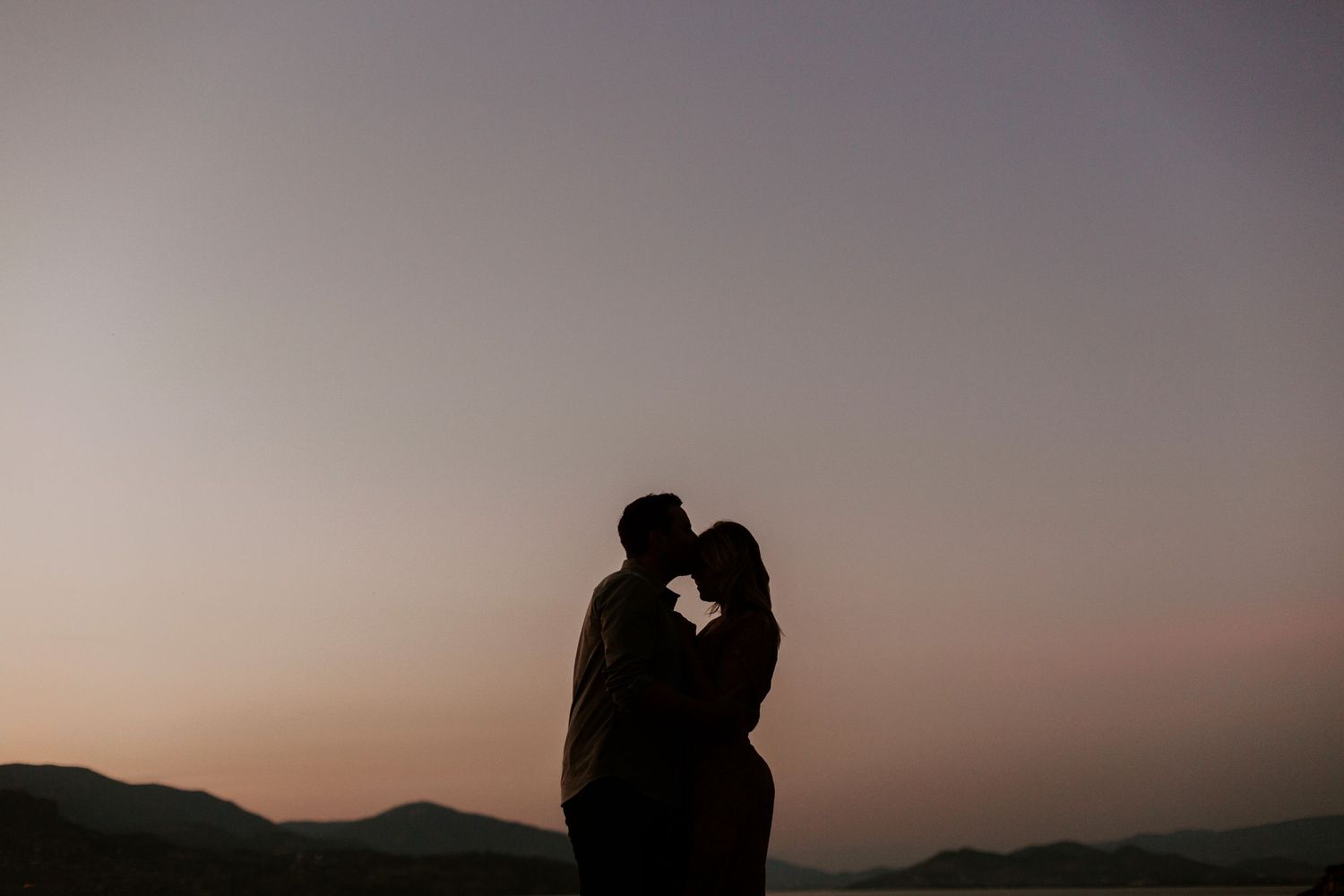 Silhouette of a romantic couple kissing at sunset with rolling hills and a colorful dusk sky in the background.