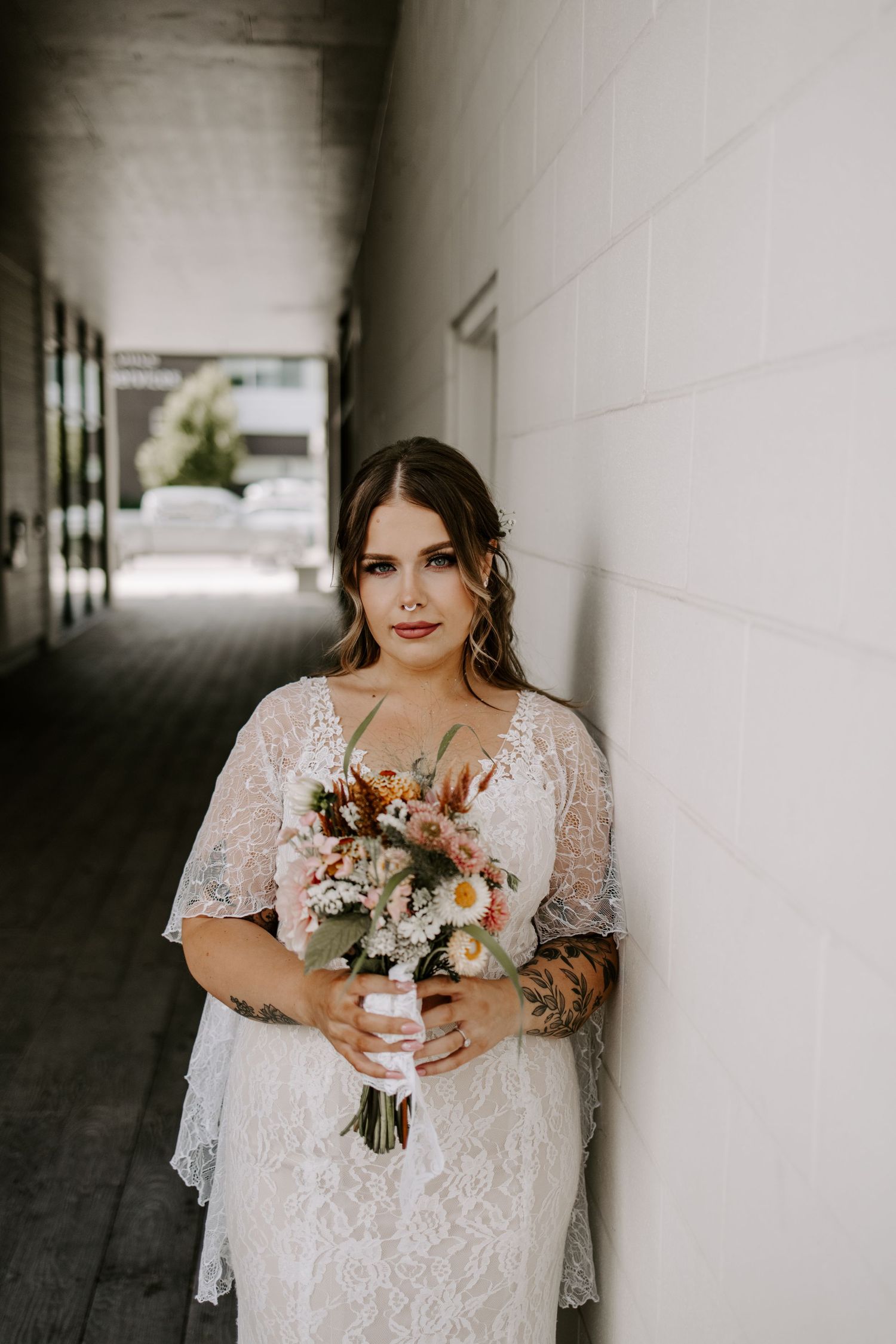 Bride in lace wedding dress holding floral bouquet with daisies, leaning against white wall in corridor.