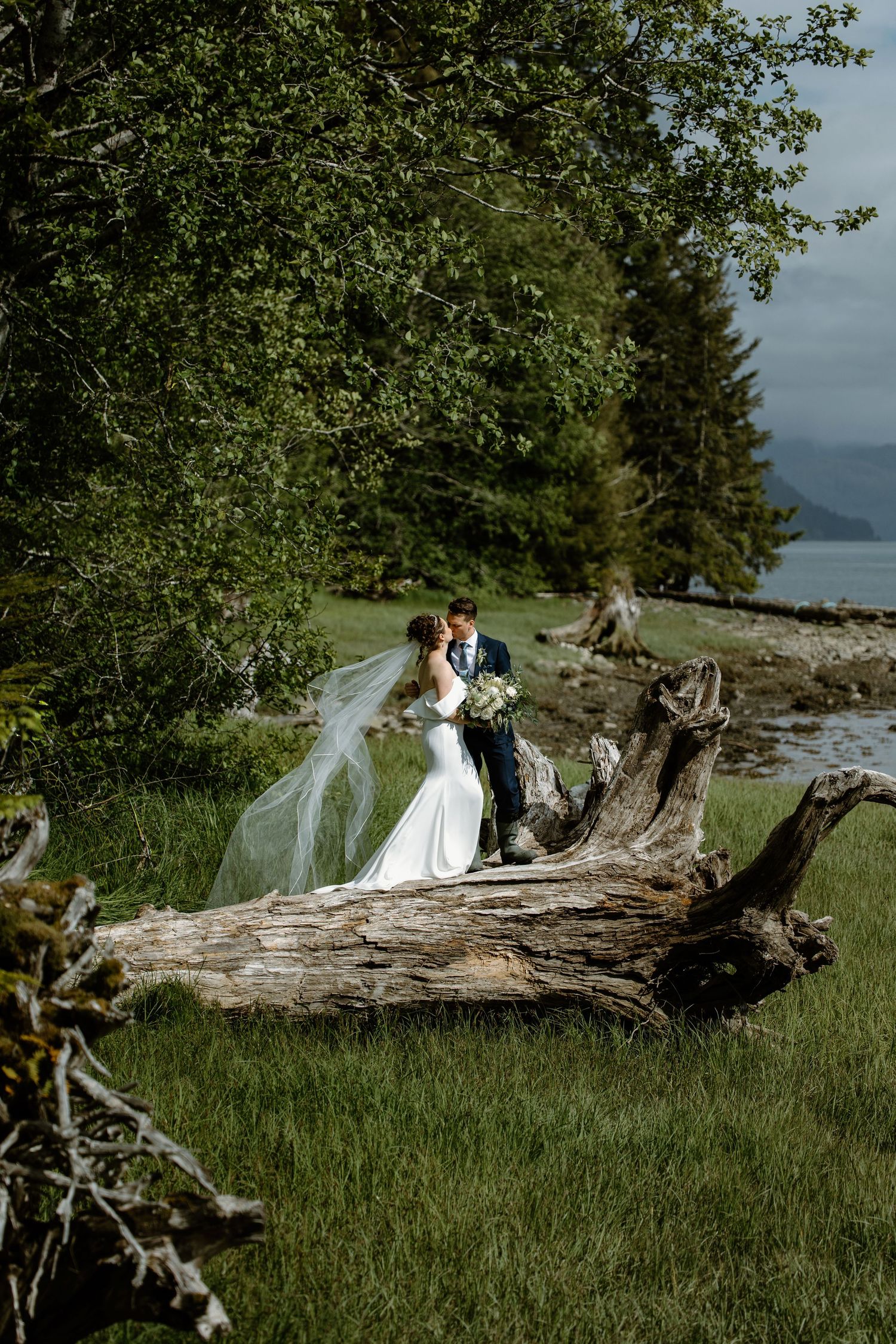 Bride and groom share a romantic kiss beside a driftwood log in a lush green waterfront setting.