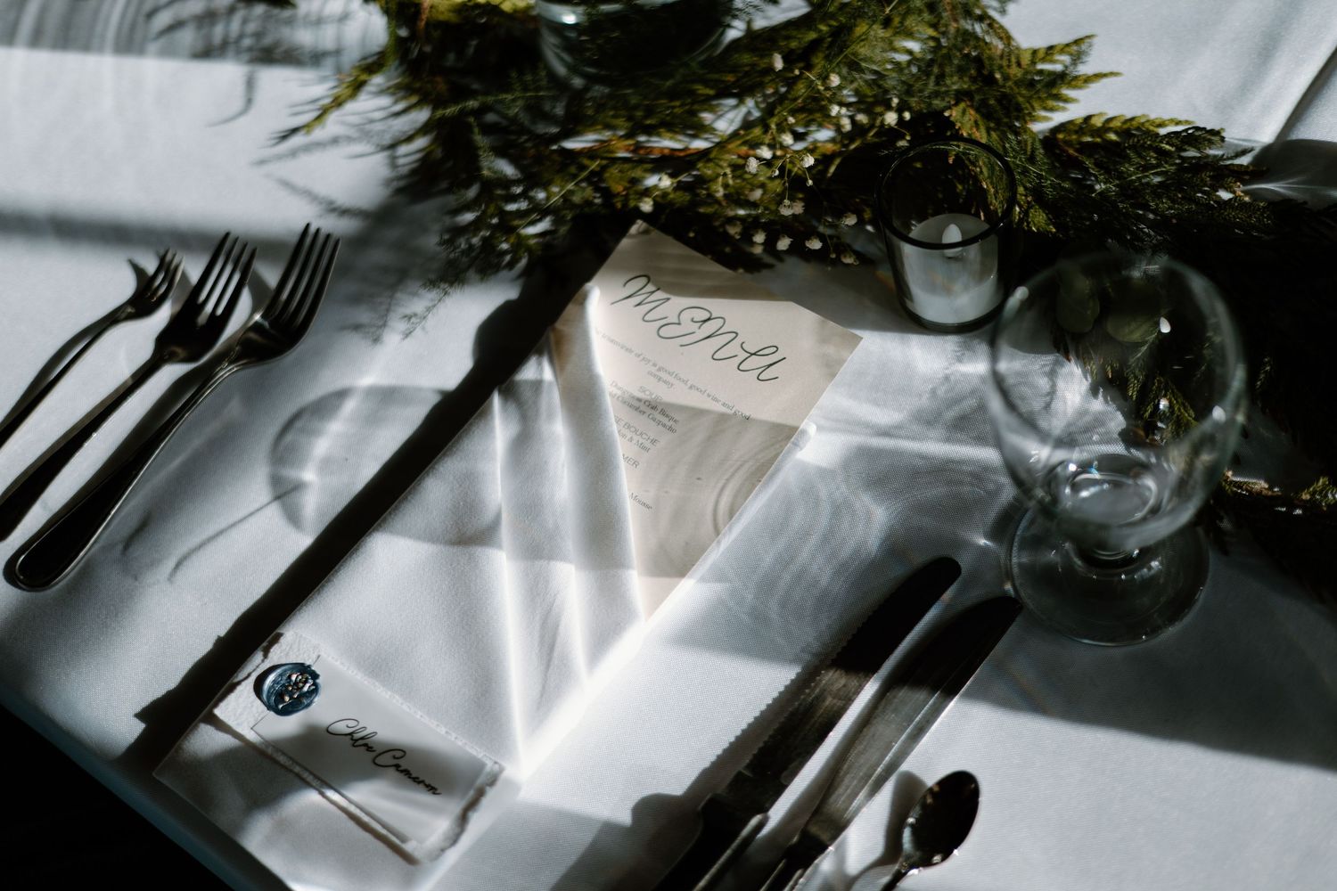 Elegant table setting with silverware, menu card and evergreen decor in dramatic lighting.