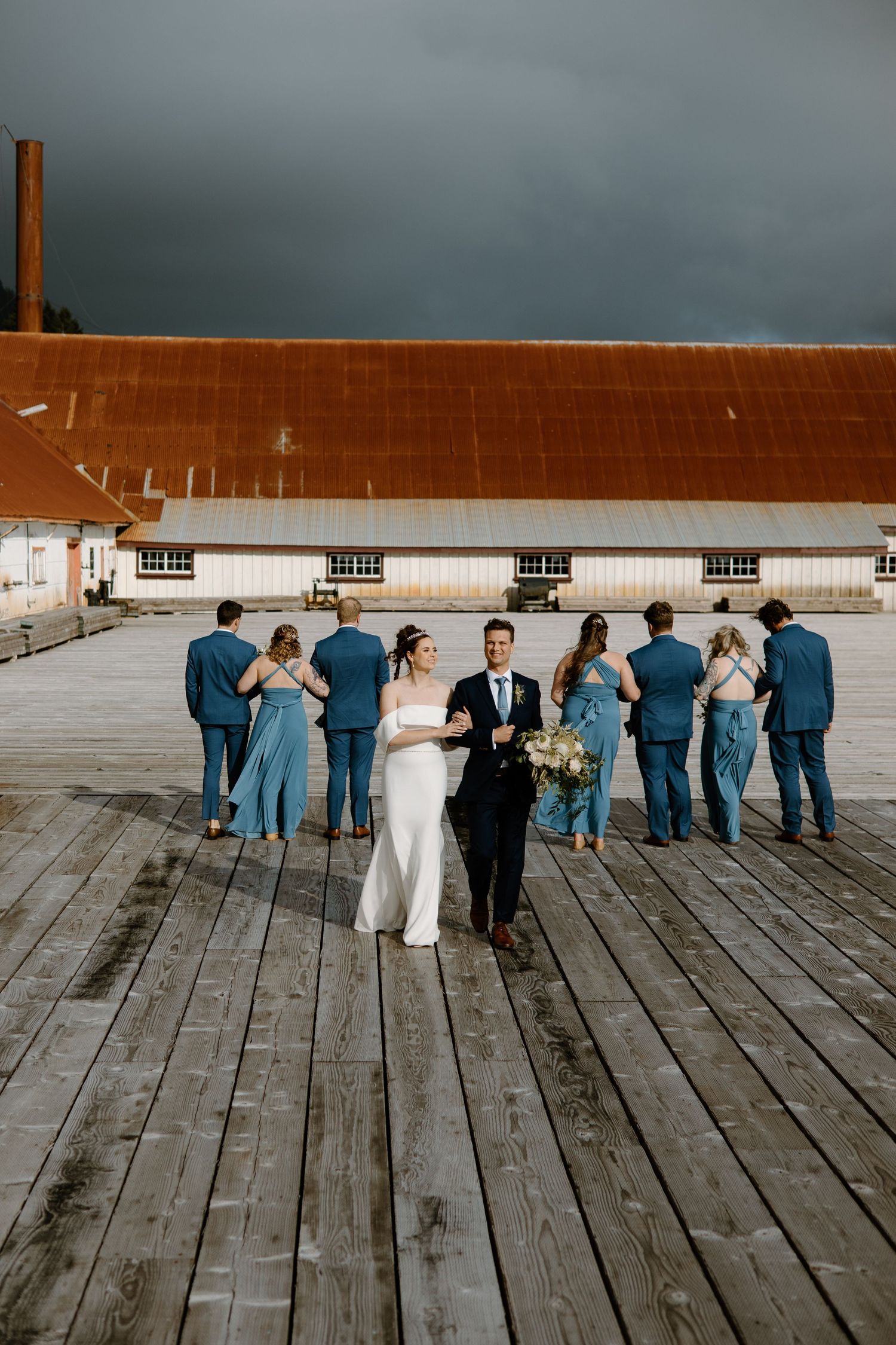 Wedding party lined up on wooden dock creating artistic silhouette against moody sky.