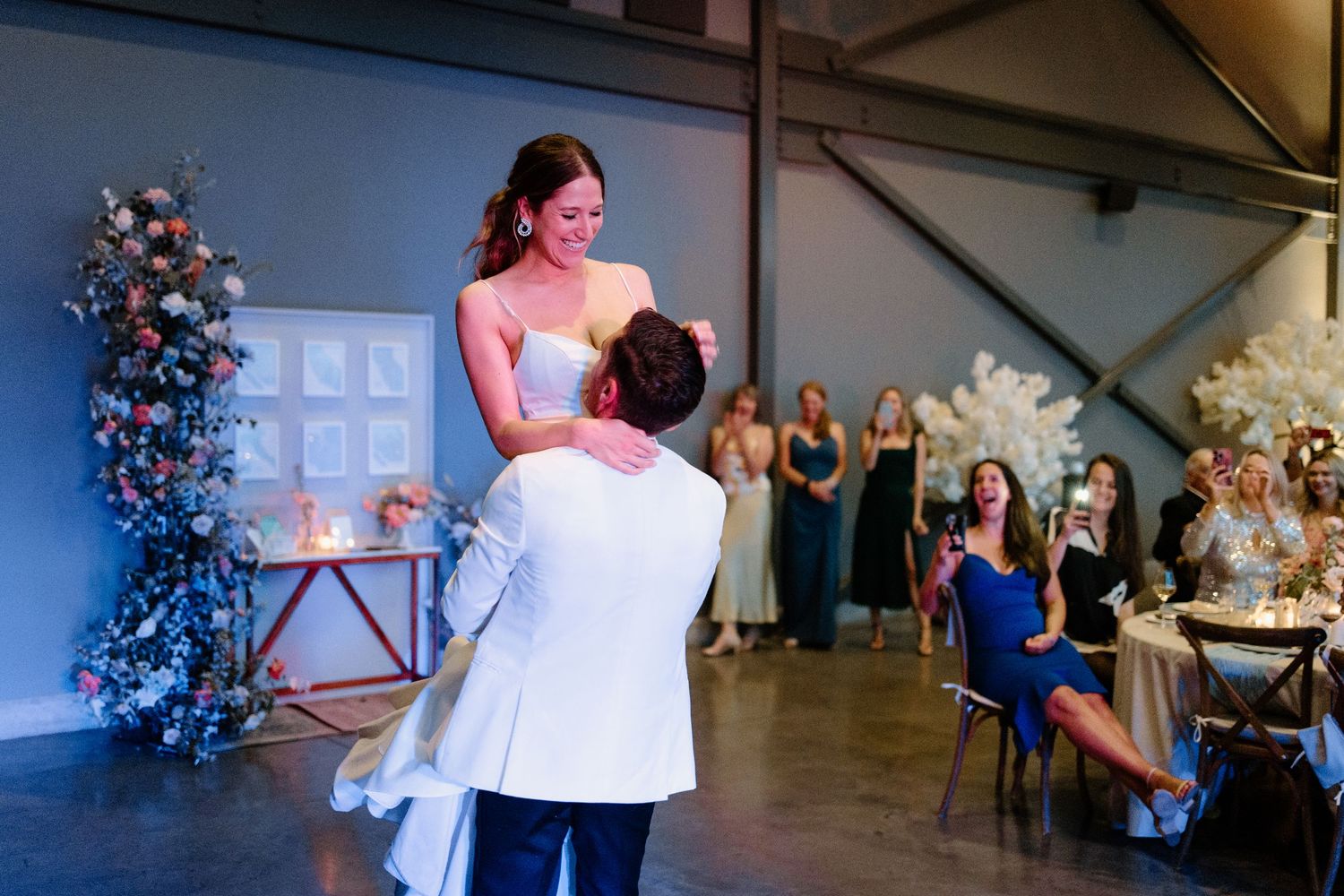 Groom in white jacket lifts bride in strapless gown during first dance at elegant wedding reception.