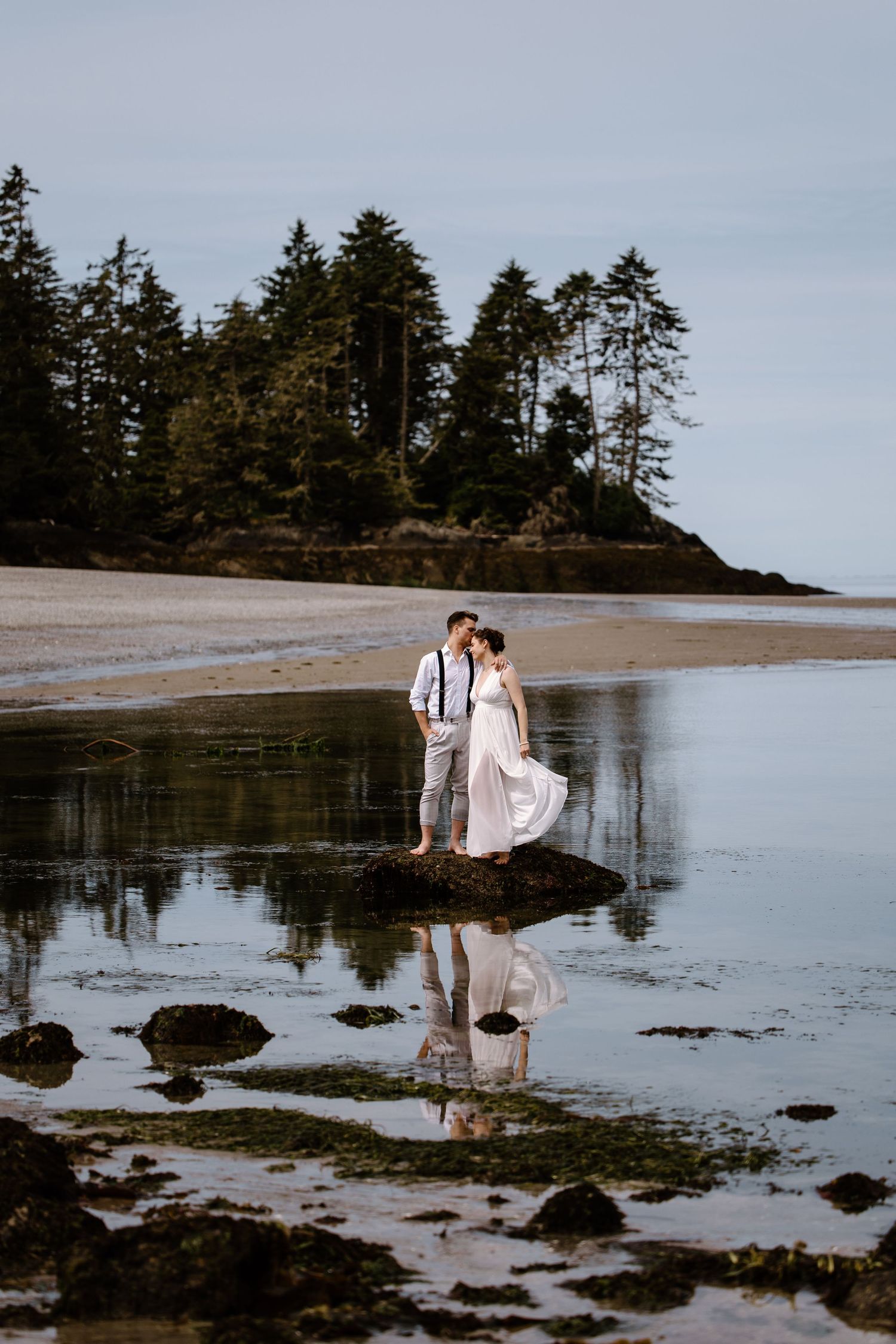 Couple in wedding attire standing on rocks at low tide beach with evergreen trees reflected in calm water.