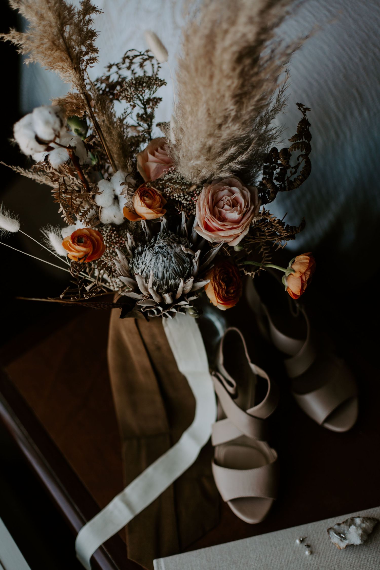 Elegant wedding shoes and floral arrangement with roses and pampas grass against dark background in moody lighting.
