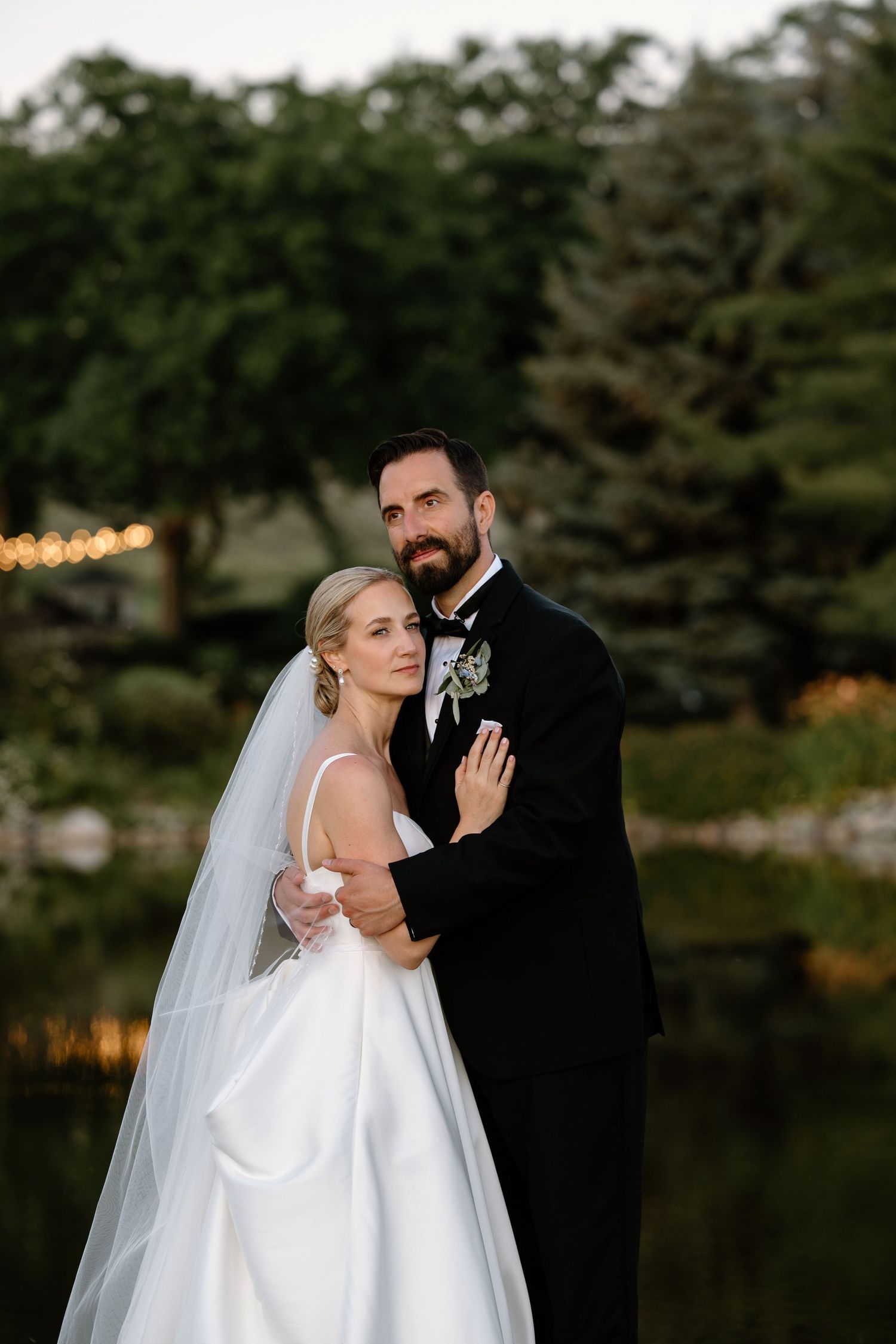 Bride in white gown and veil embraces groom in black tuxedo outdoors near a pond with lush green trees.