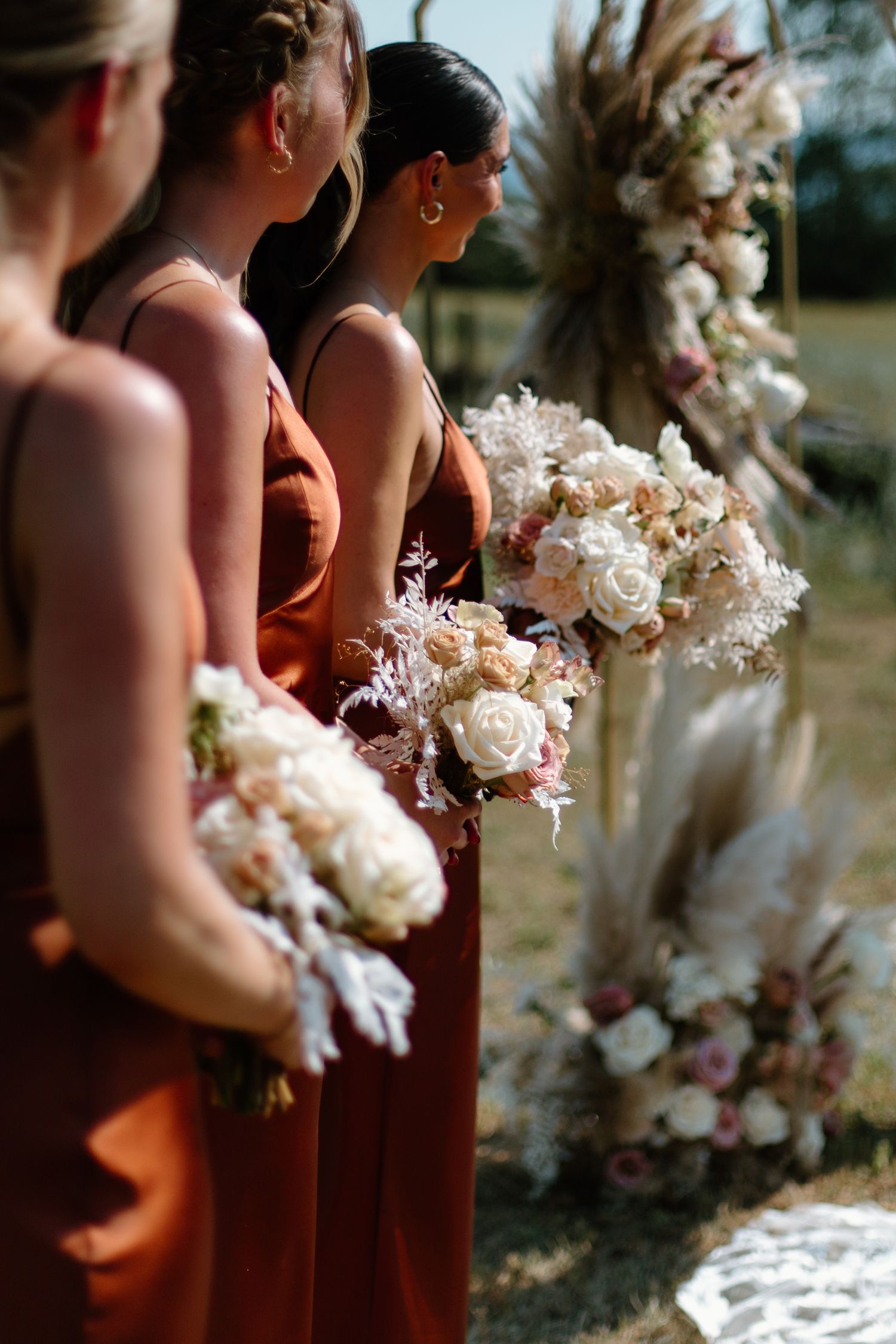 Bridesmaids in terracotta dresses hold elegant bouquets of white roses and dried flowers at an outdoor wedding ceremony.