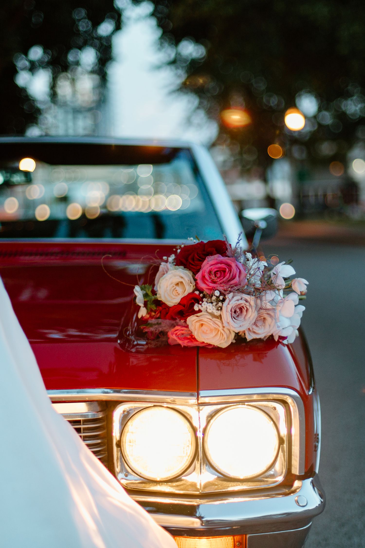 A close-up view of vintage car headlights with pink and white roses decorating the front of a classic red automobile.