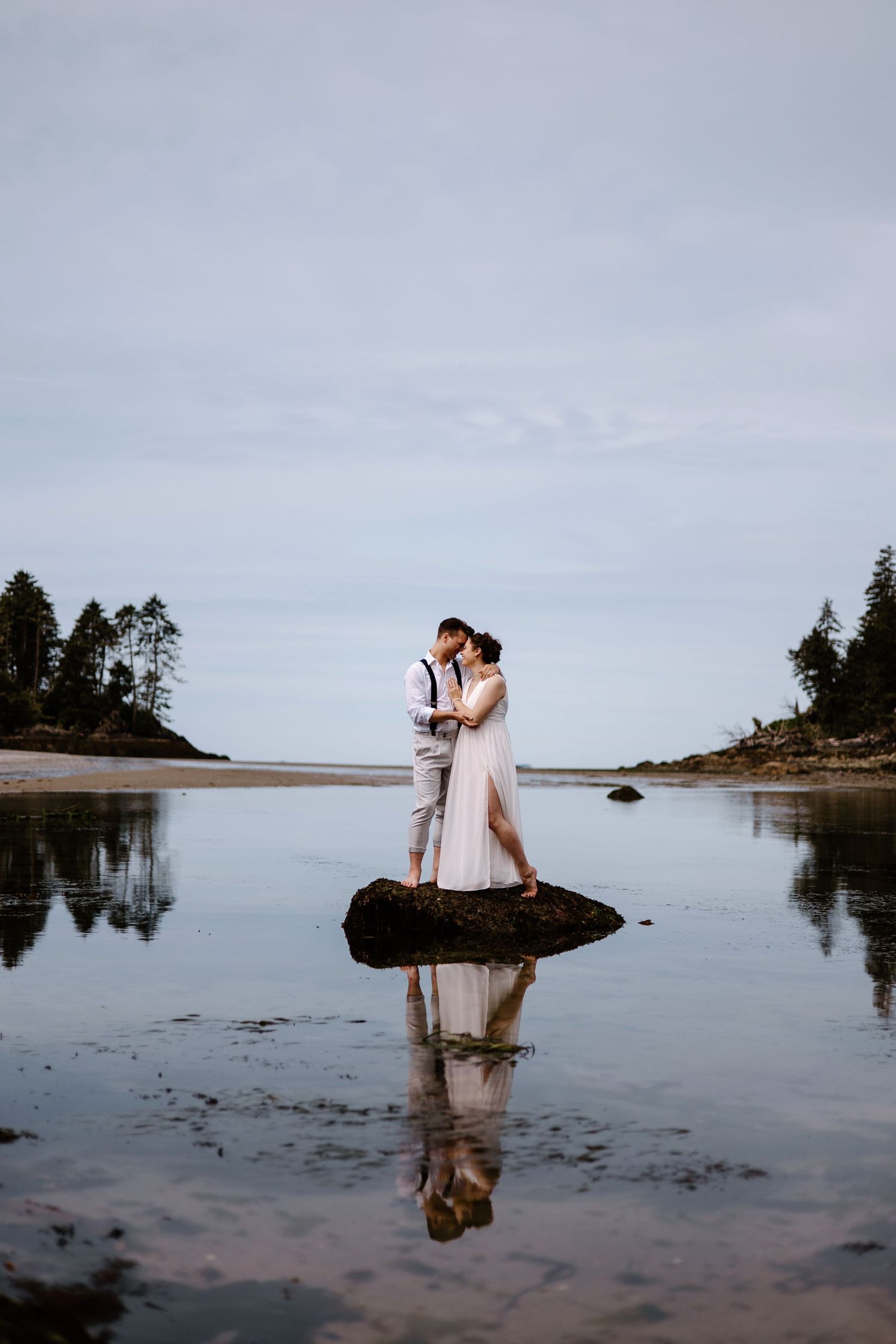 Wedding couple reflected in tidal pools at sunset with evergreen coastline.