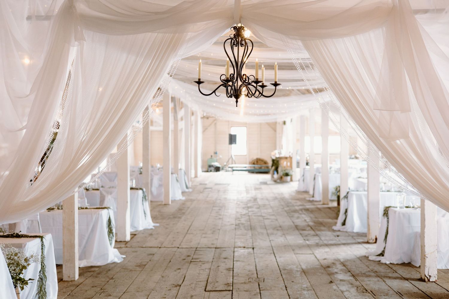 White sheer fabric drapes line a rustic wooden hall with an elegant chandelier creating a romantic wedding venue atmosphere.