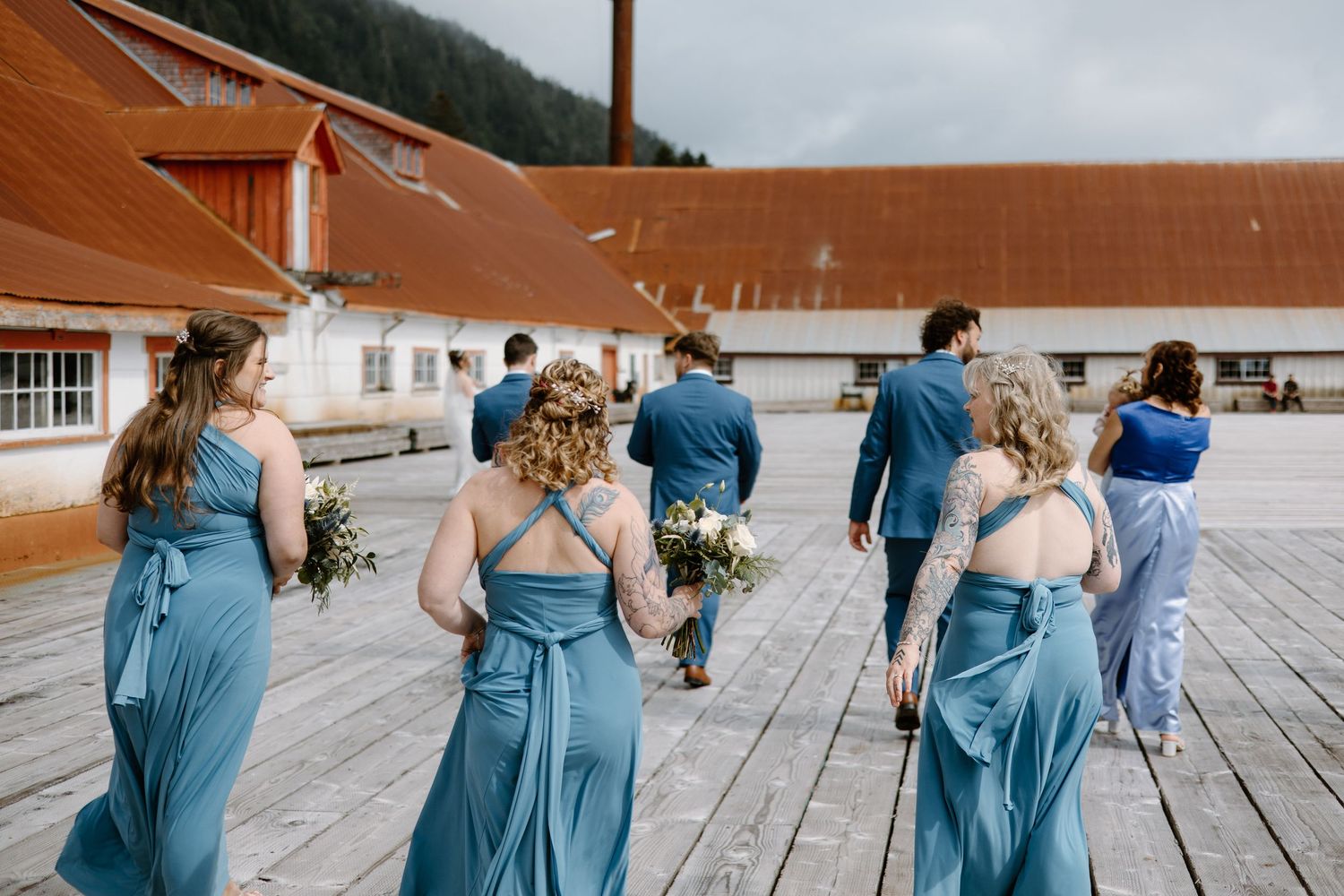Bridesmaids in blue dresses walking along wooden dock during wedding photoshoot.