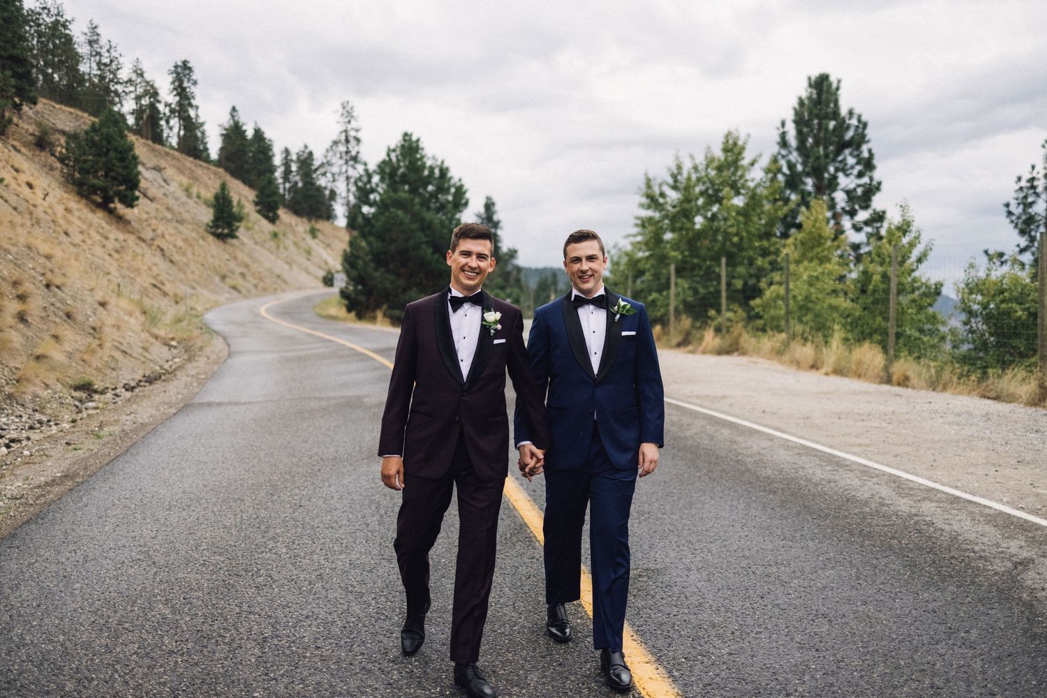 Two grooms in tuxedos hold hands on a winding mountain road surrounded by evergreen trees on their wedding day.