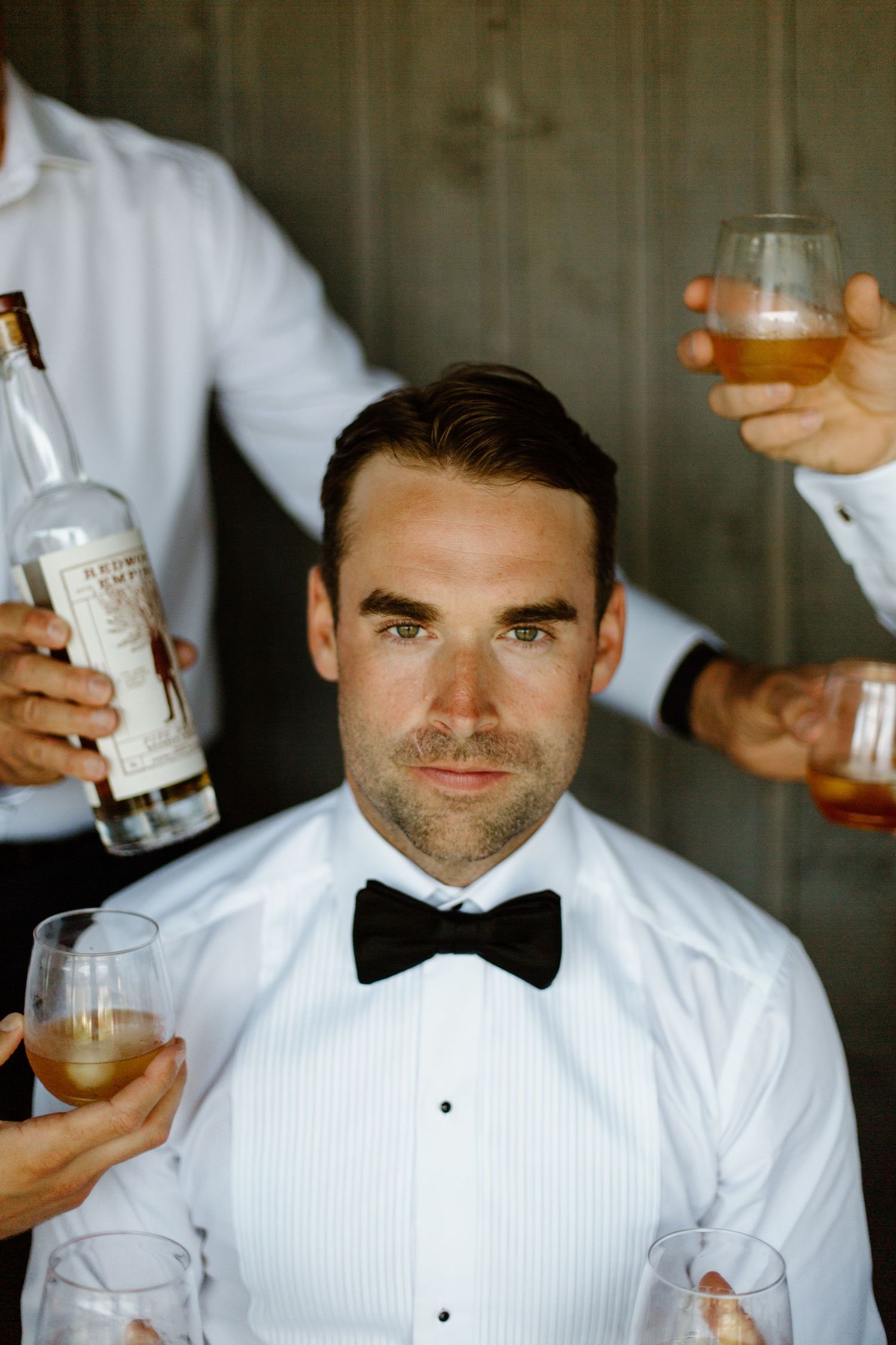 Groom in white tuxedo shirt and bow tie surrounded by hands holding bourbon bottle and cocktail glasses getting ready on his wedding day.