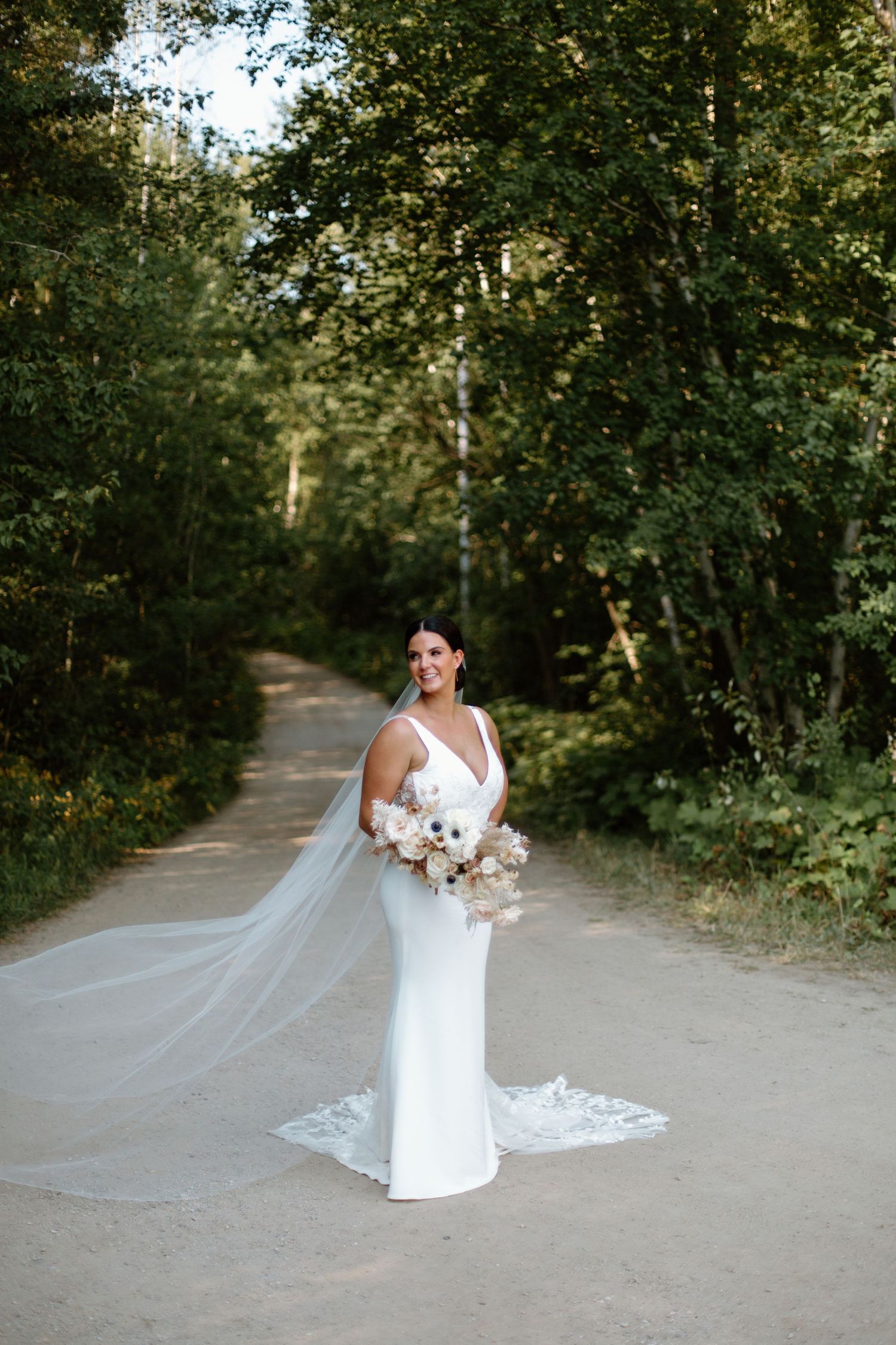 A bride in a white fitted gown and flowing veil stands on a forest path holding a white floral bouquet.