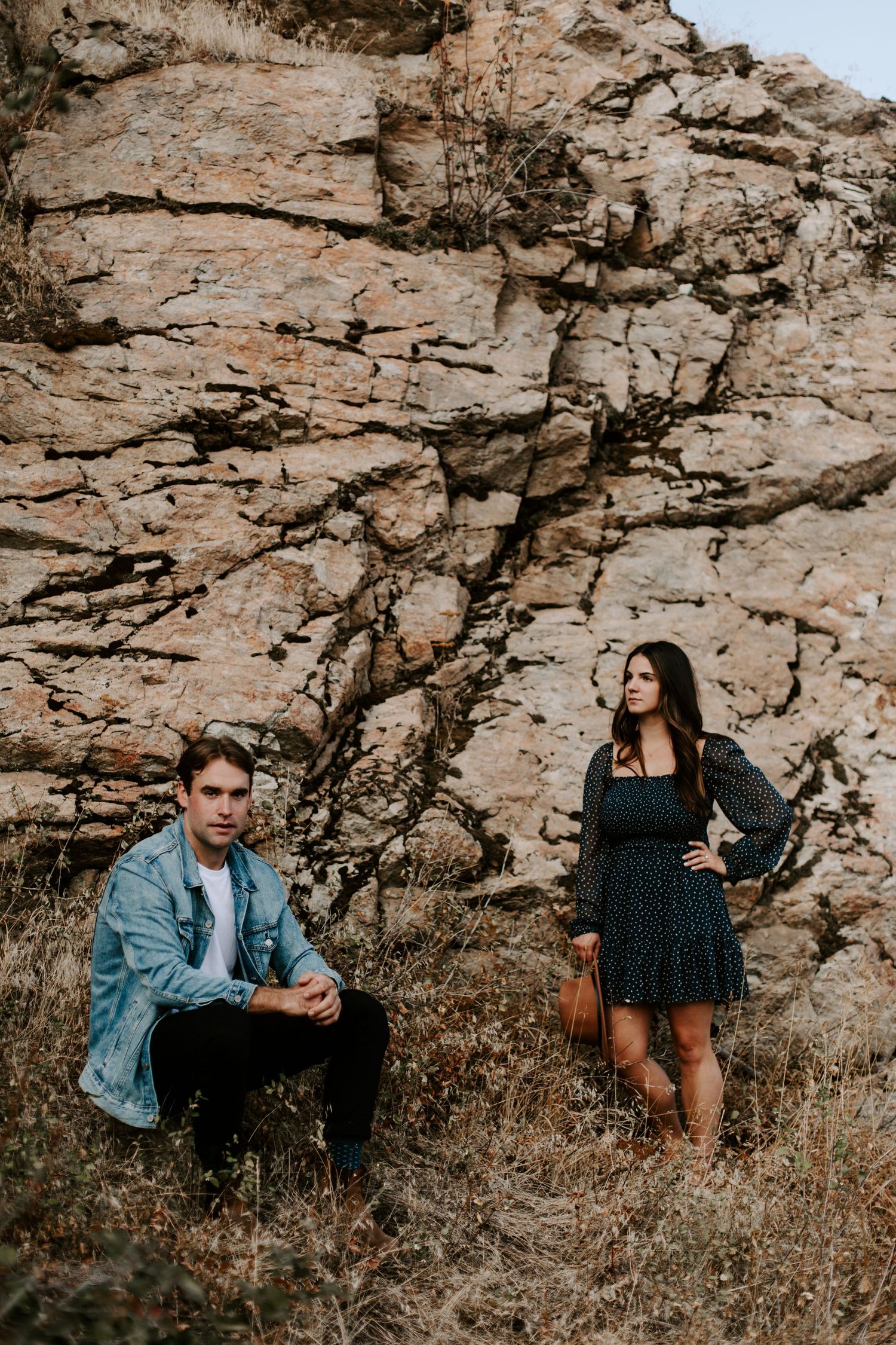 A man in a denim jacket and a woman in a floral dress pose against a rocky cliff backdrop outdoors.