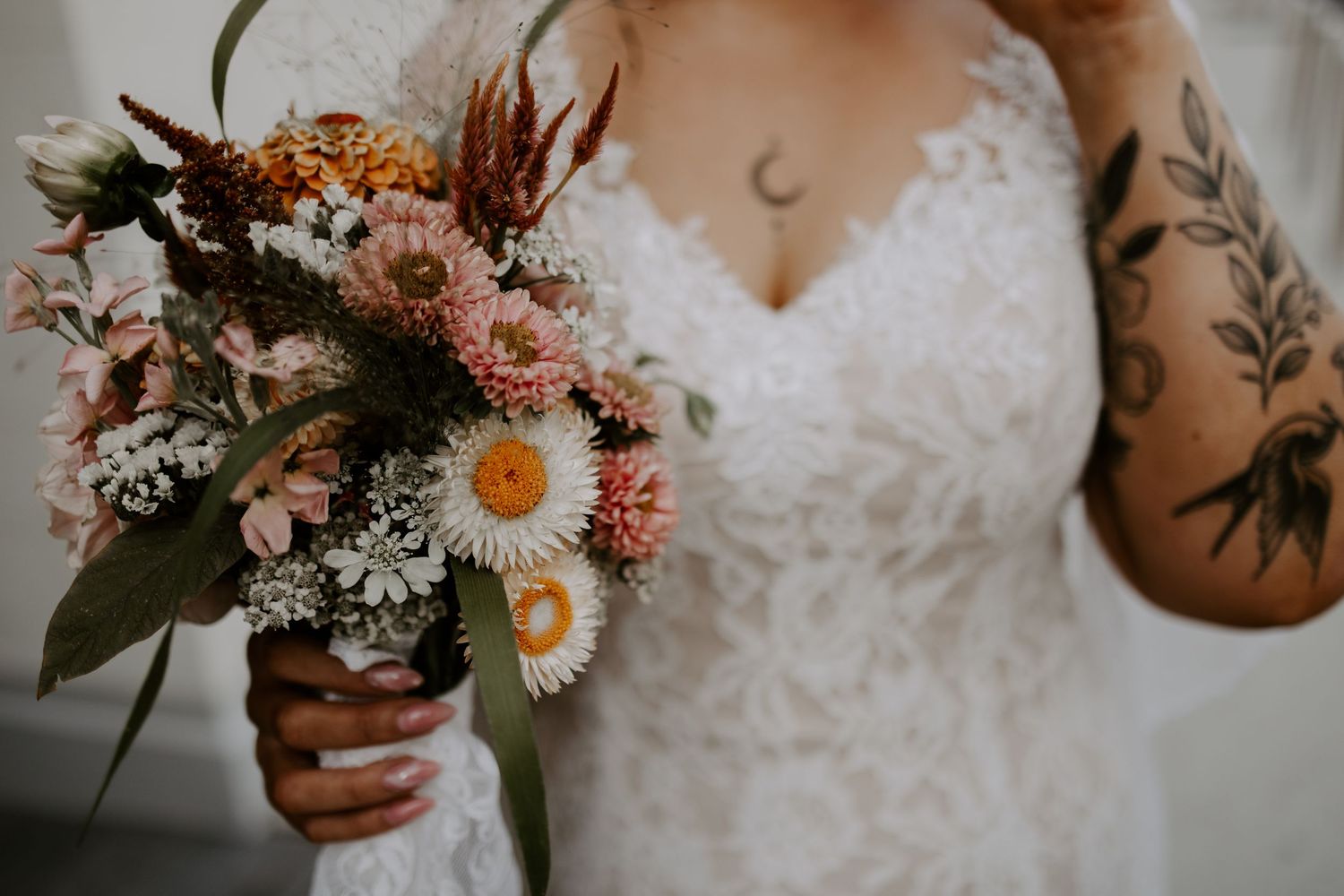 Close-up of a wedding bouquet with daisies and dried flowers held against a lace dress with visible tattoos.