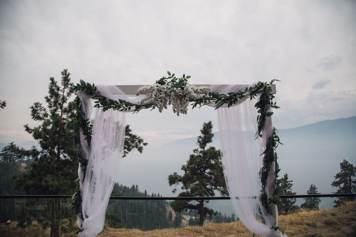 A wedding arch decorated with white fabric and greenery stands against a scenic mountain landscape.