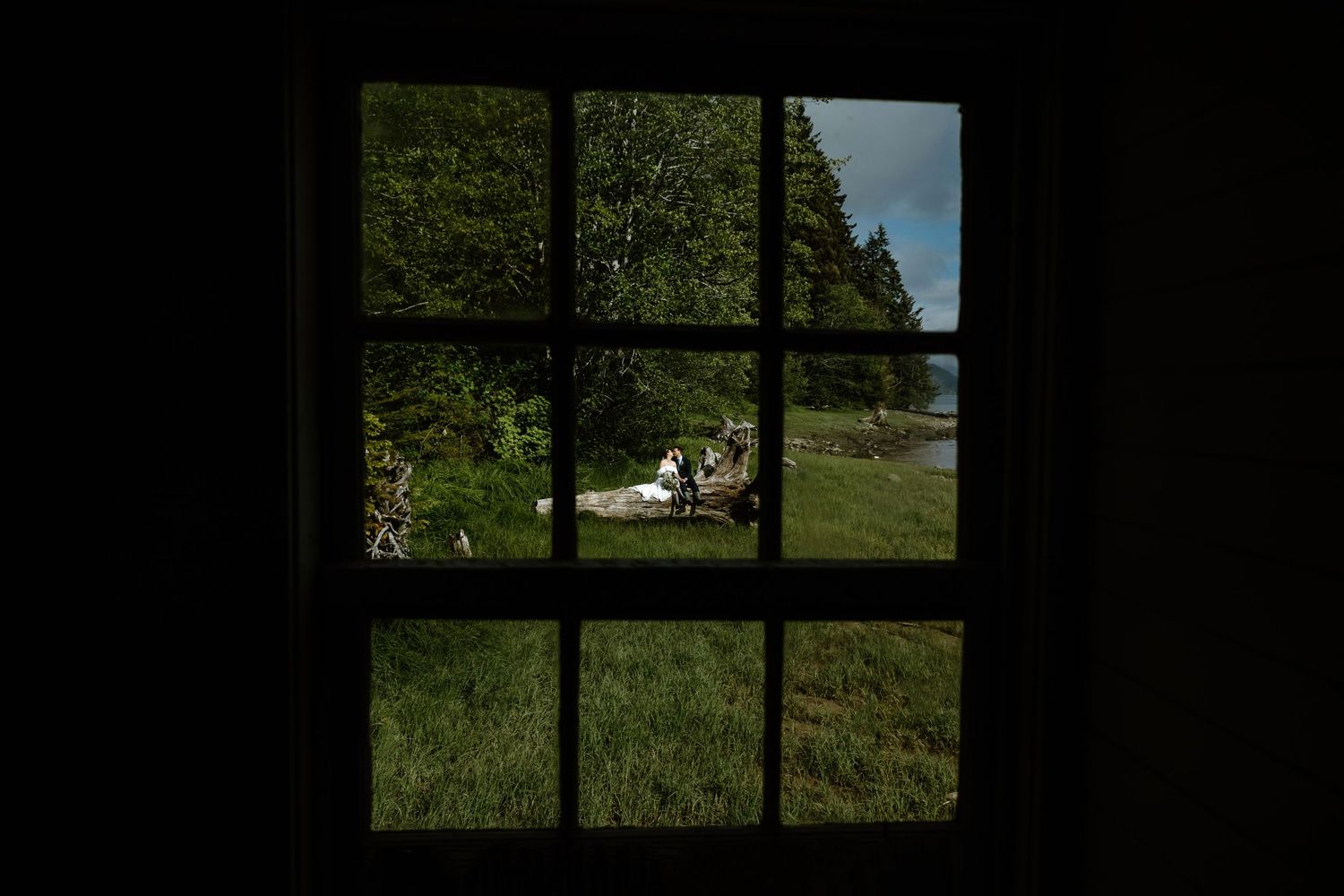 View through a dark window frame of people relaxing on a bench surrounded by lush green trees and grass.