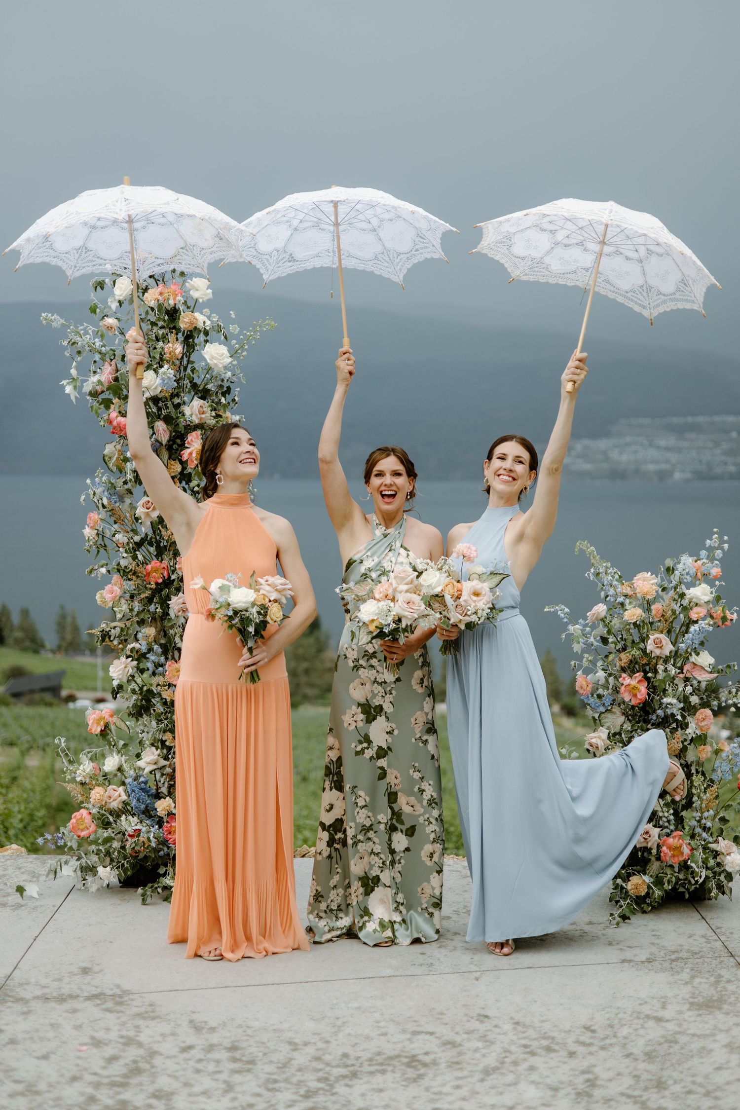 Three bridesmaids in pastel gowns holding white lace umbrellas and floral bouquets at an outdoor wedding ceremony.