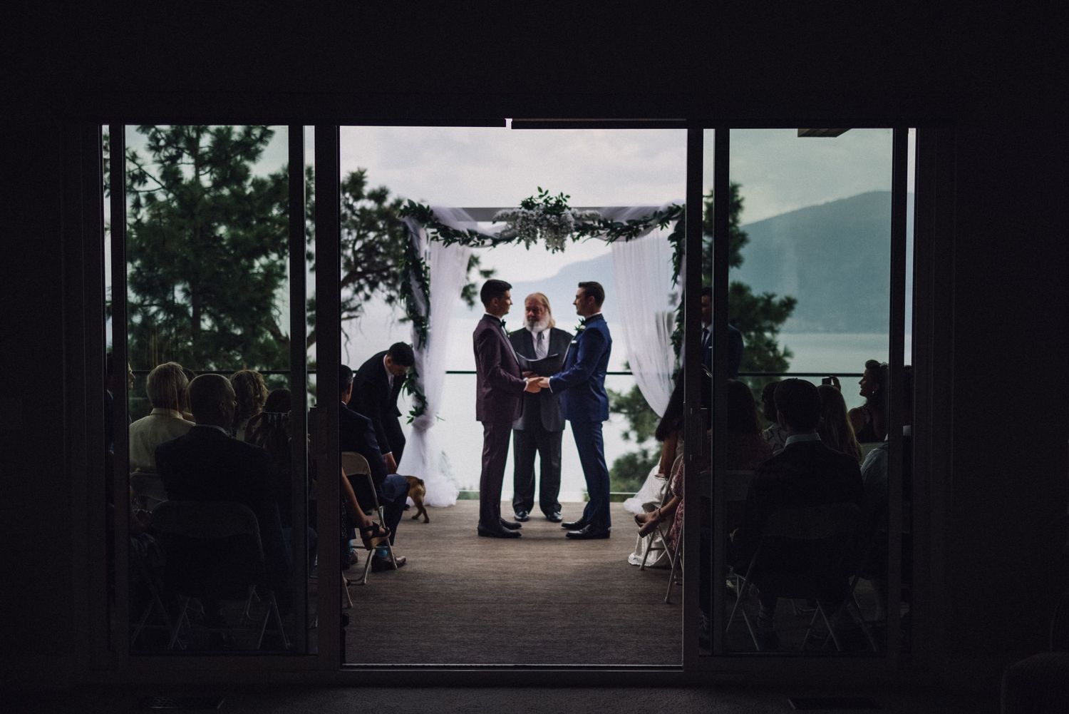 Two grooms exchange vows at an outdoor wedding ceremony framed by an elegant floral arch with lake views.