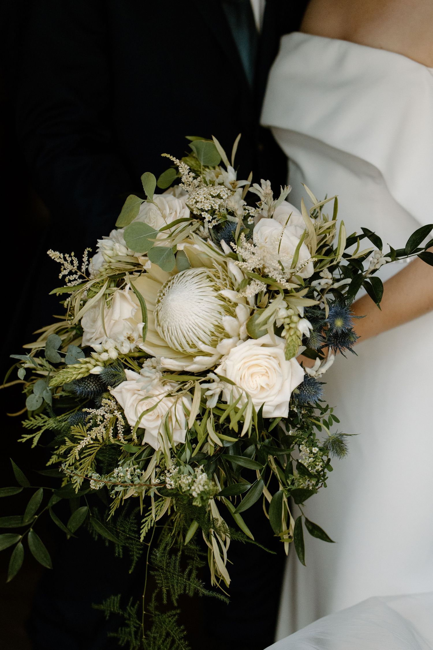 Bridal bouquet featuring white king protea, roses, eucalyptus, and blue thistle held by bride in white gown.