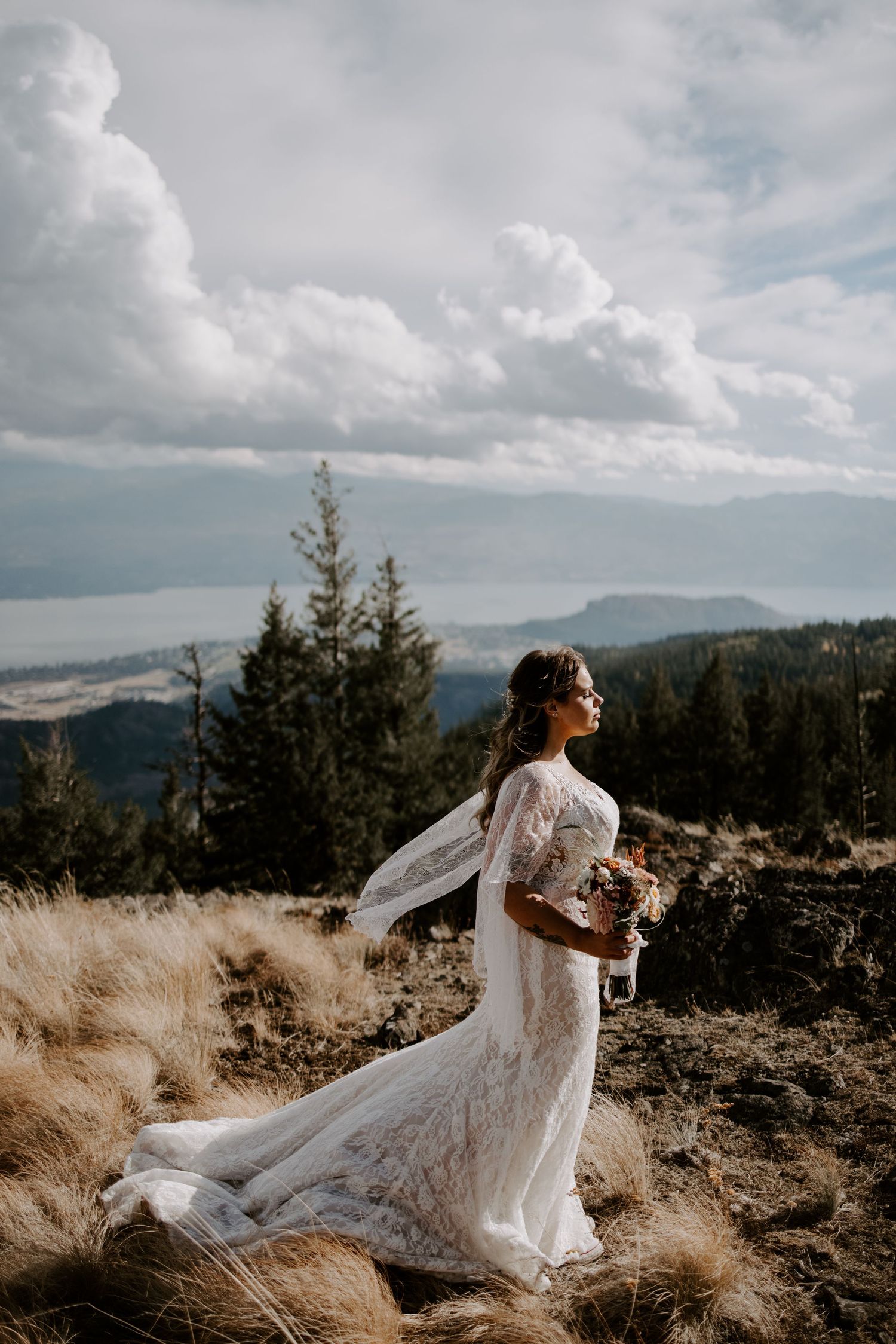 Bride's white dress and veil flow dramatically in the wind on mountain summit with evergreen trees below.