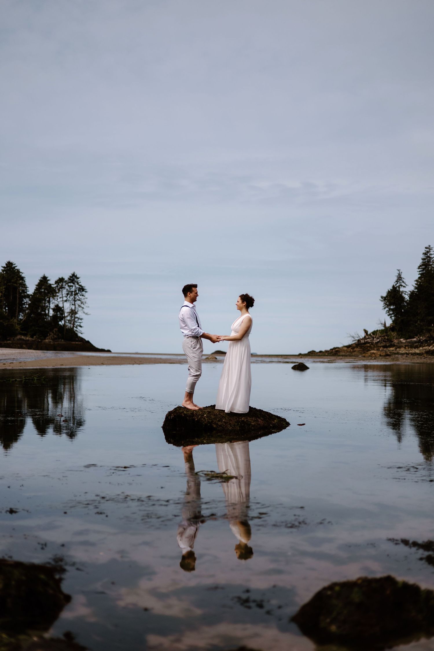 Two figures stand together on a tranquil rocky shoreline at dusk with reflections in the calm water.