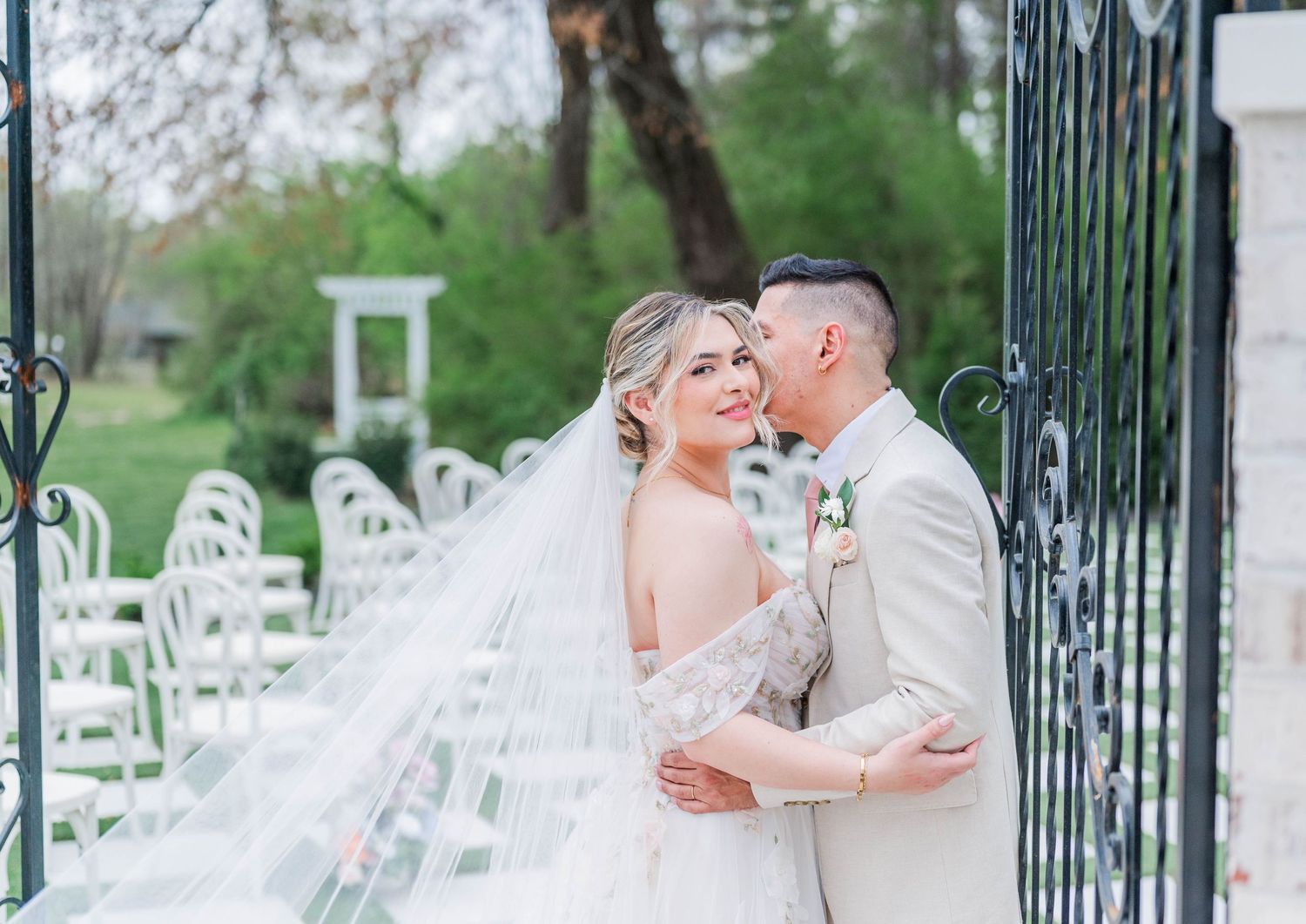 A couple shares a romantic moment by iron gates on their wedding day, with white chairs and greenery in the background.