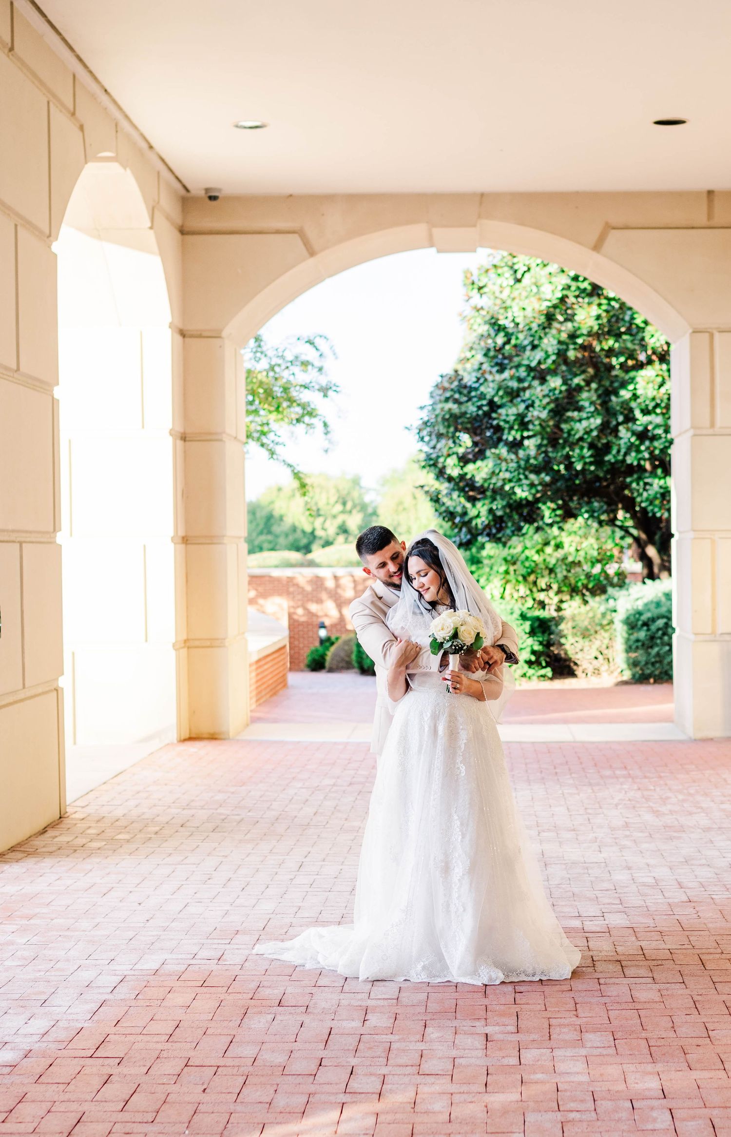 A romantic wedding couple embraces under an elegant arched walkway with brick pavers and lush greenery in the background.