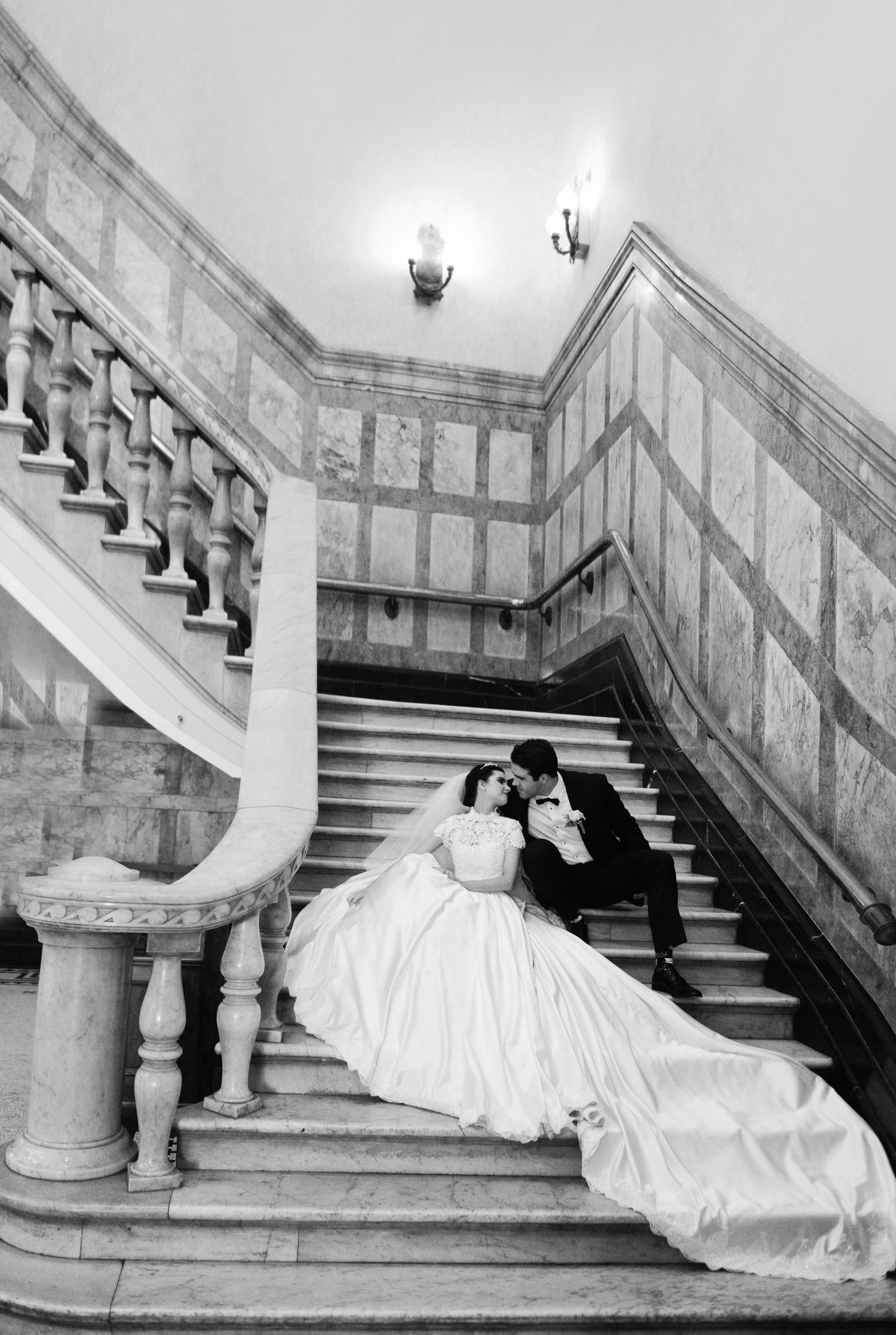 Black and white photo of bride and groom sitting on grand marble staircase in elegant wedding portrait.