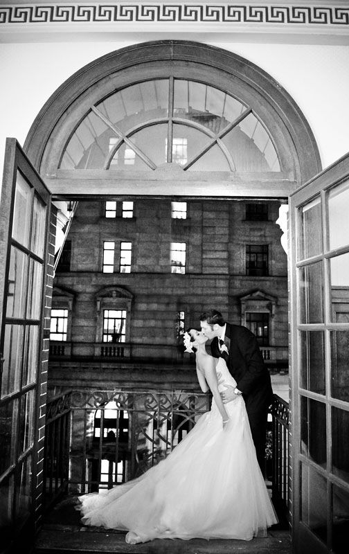 Black and white wedding photo of bride and groom kissing on balcony framed by elegant arched doorway.
