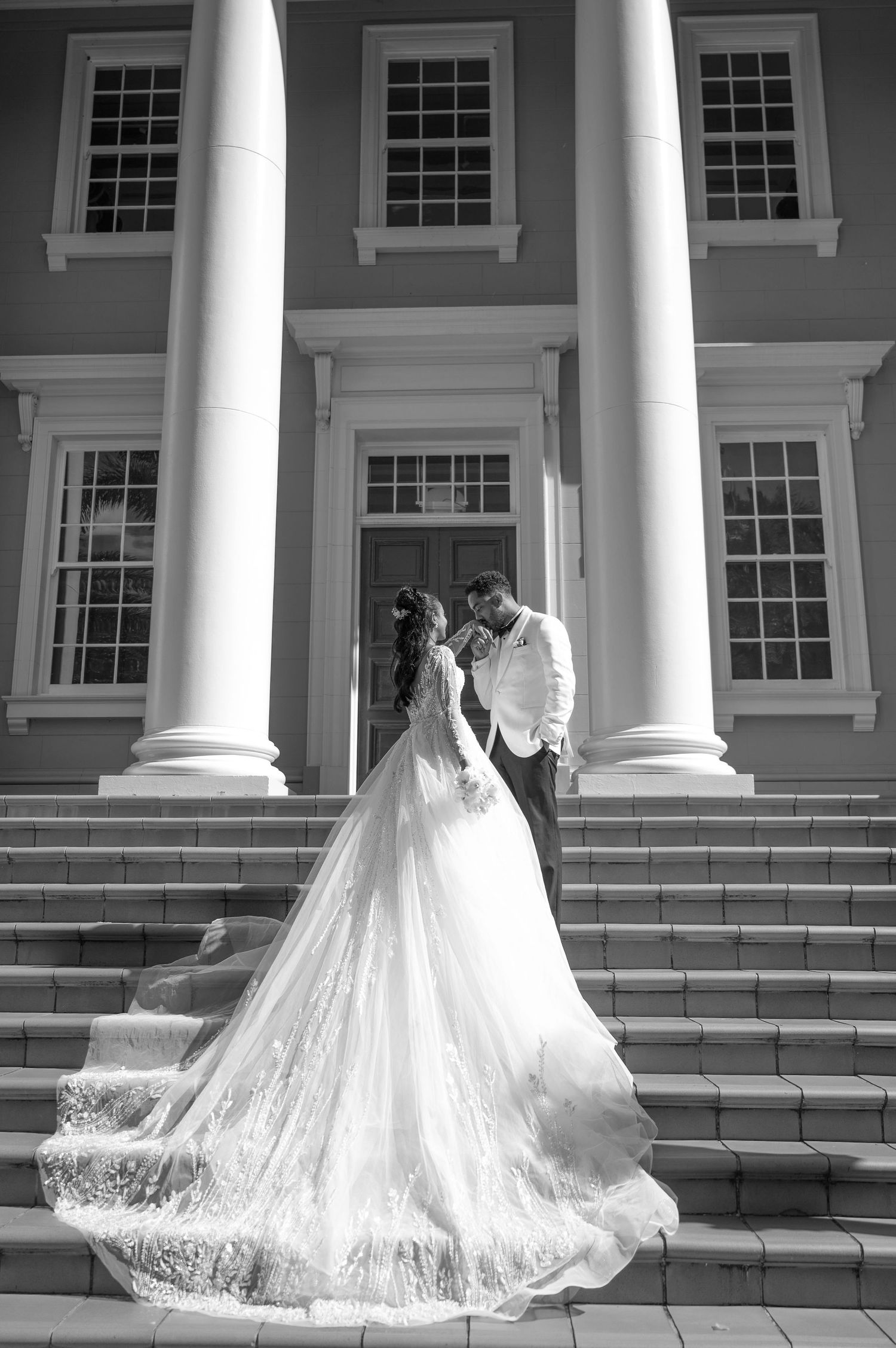 Black and white photo of bride and groom embracing on grand staircase of elegant columned mansion.