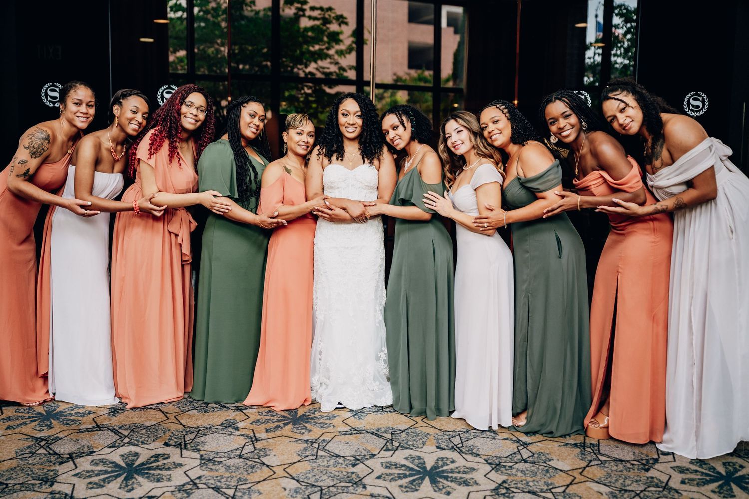 Wedding party in coral and sage green dresses pose together in a curved line formation outdoors.