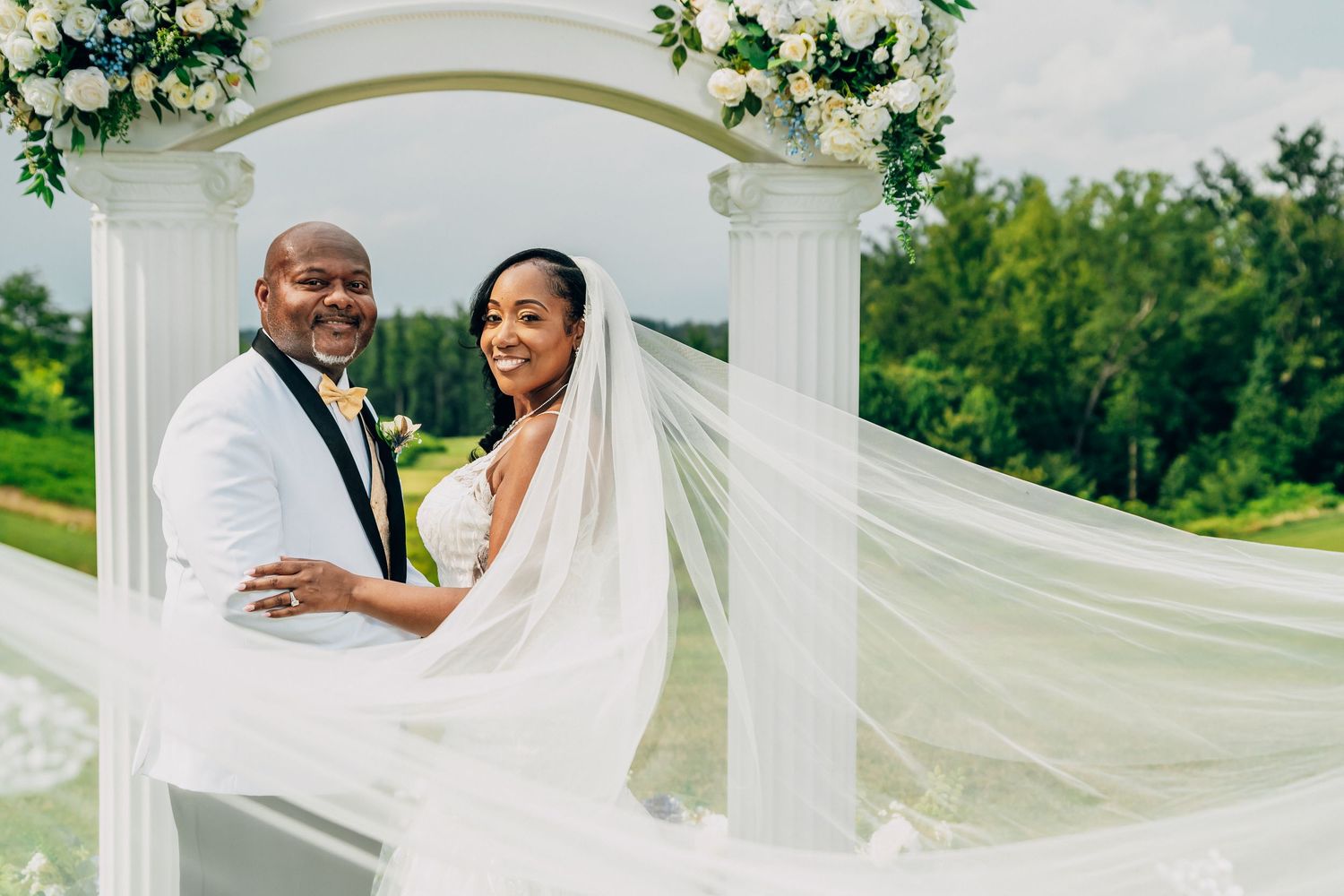 A joyful couple shares a tender moment under a white wedding arch decorated with greenery as the bride's veil flows in the breeze.