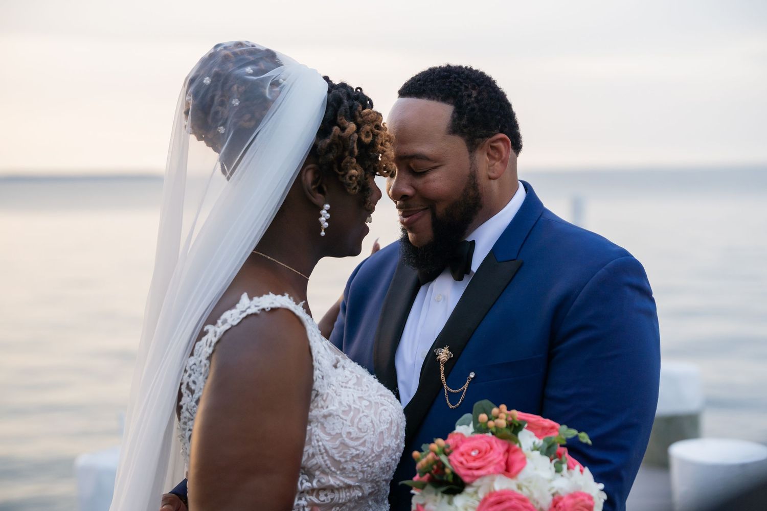 Romantic beachside wedding couple sharing an intimate moment at sunset with pink bouquet and navy suit.