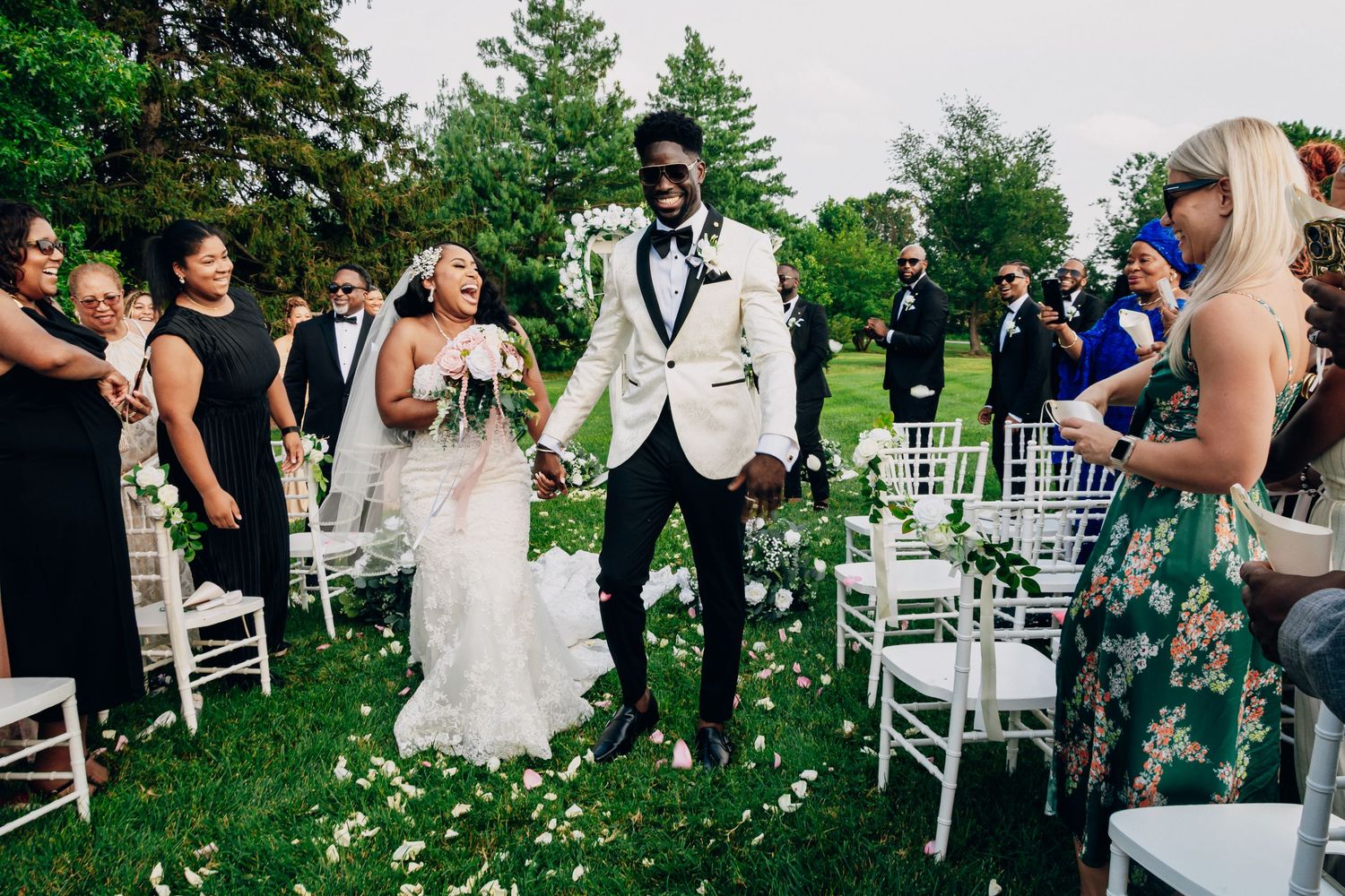 A wedding ceremony takes place in a lush outdoor garden setting with white chairs arranged in rows for guests.
