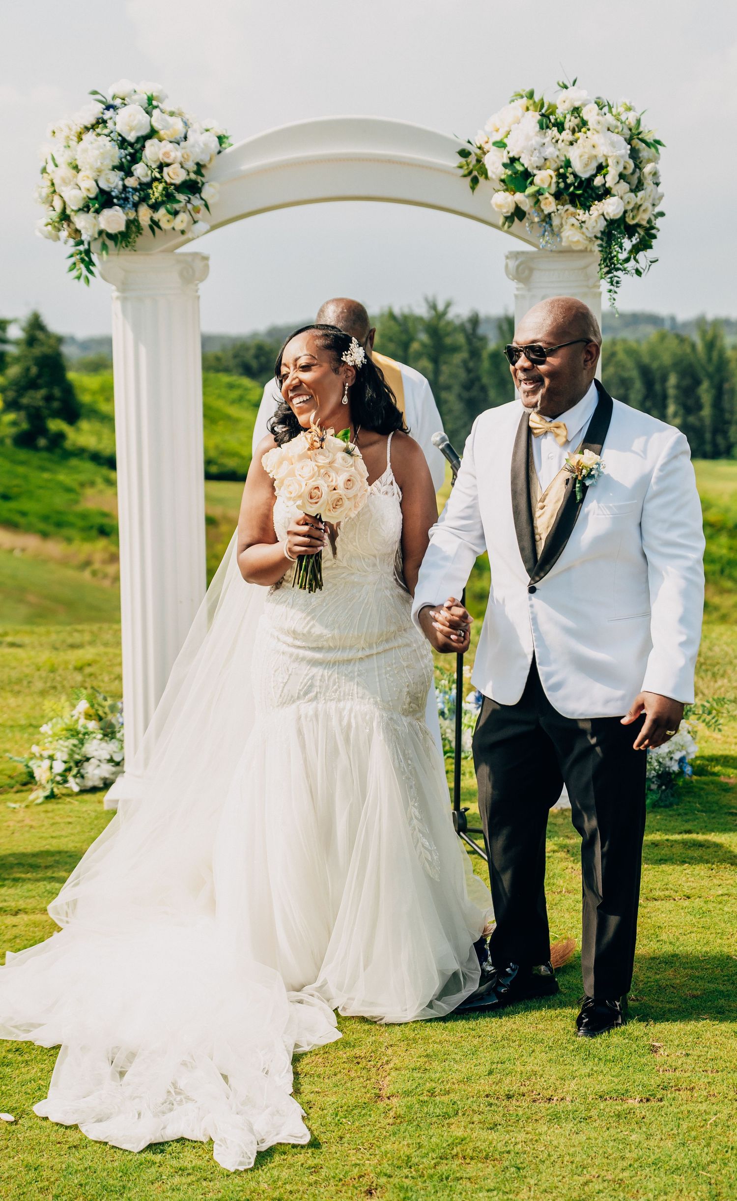 A joyful wedding ceremony under a white arch decorated with floral arrangements on a scenic hilltop.