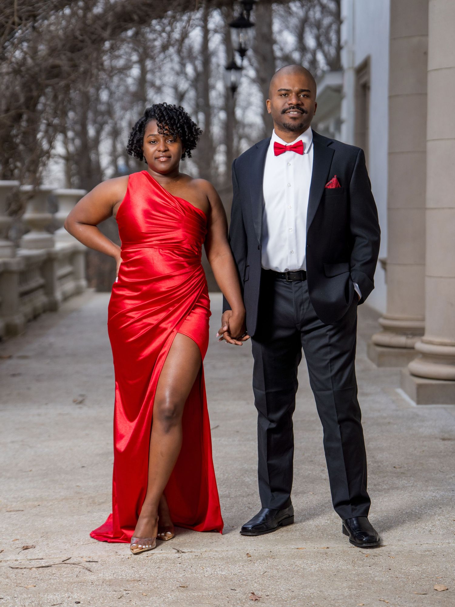 A couple in formal attire poses in an outdoor colonnade, with the woman wearing a flowing red gown and man in black tuxedo.