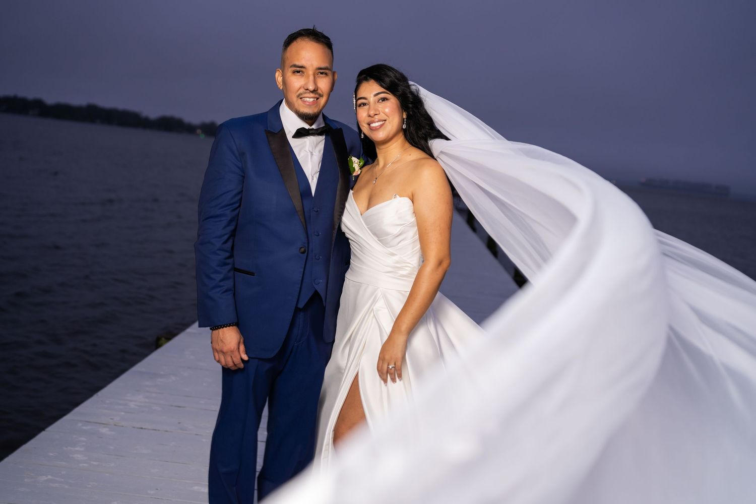 A couple poses for romantic wedding photos at sunset by the ocean with the bride's white dress flowing in the breeze.