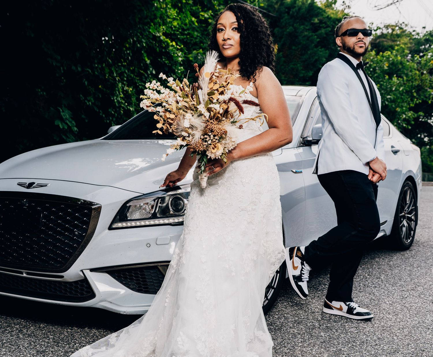 A bride in a white gown holds a dried floral bouquet while standing next to a white luxury vehicle on their wedding day.