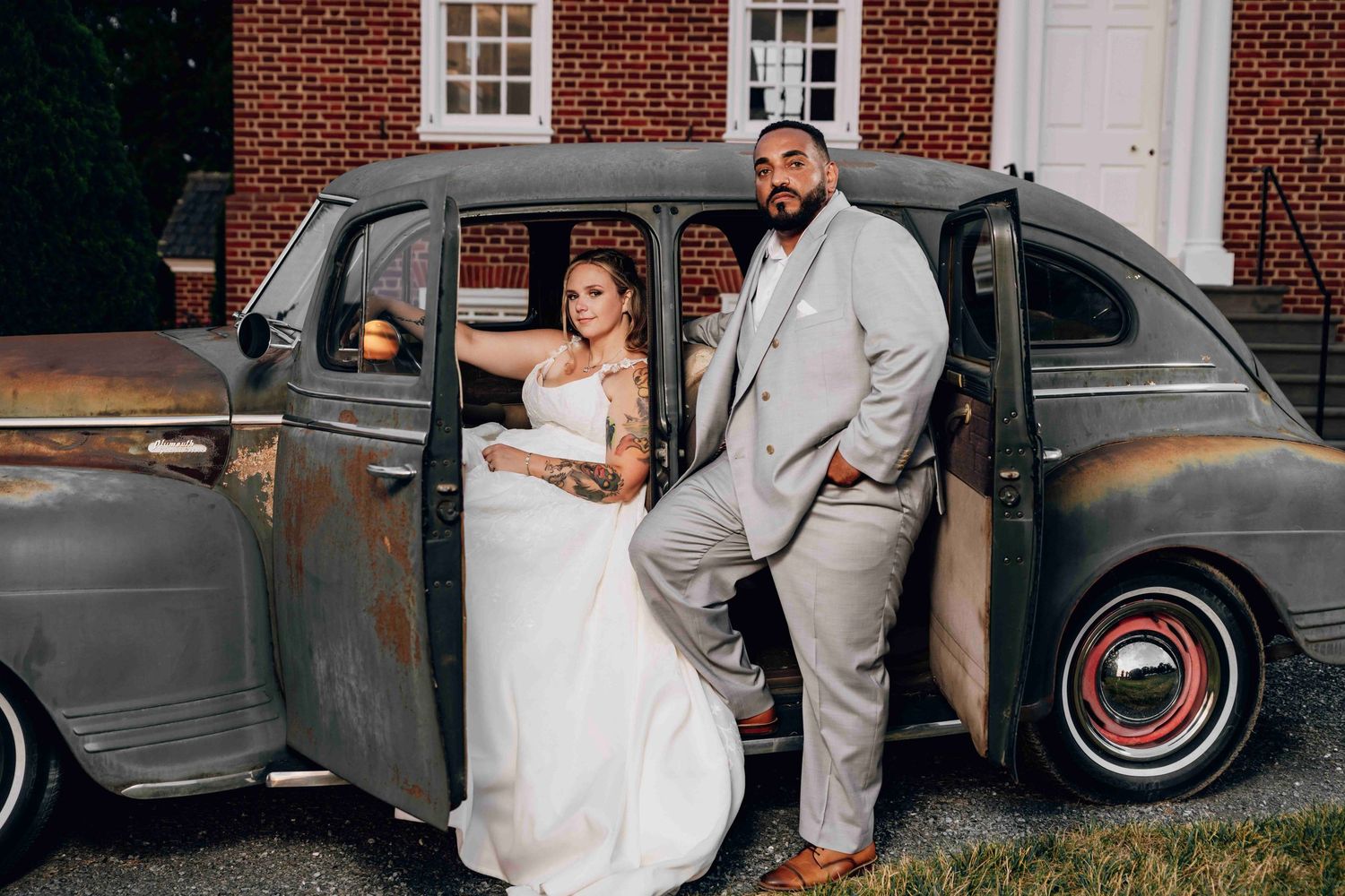 A couple poses beside a classic vintage grey automobile at their outdoor wedding celebration.
