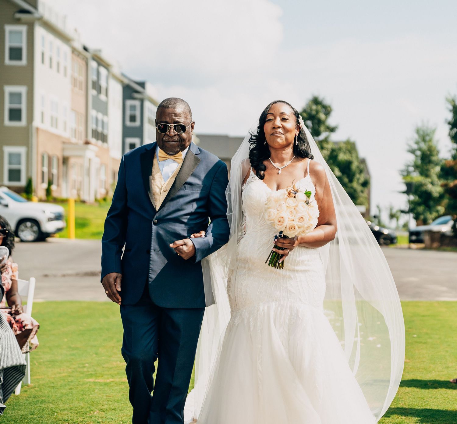 A joyful wedding couple walks together on a sunny day across a green lawn outside their venue.